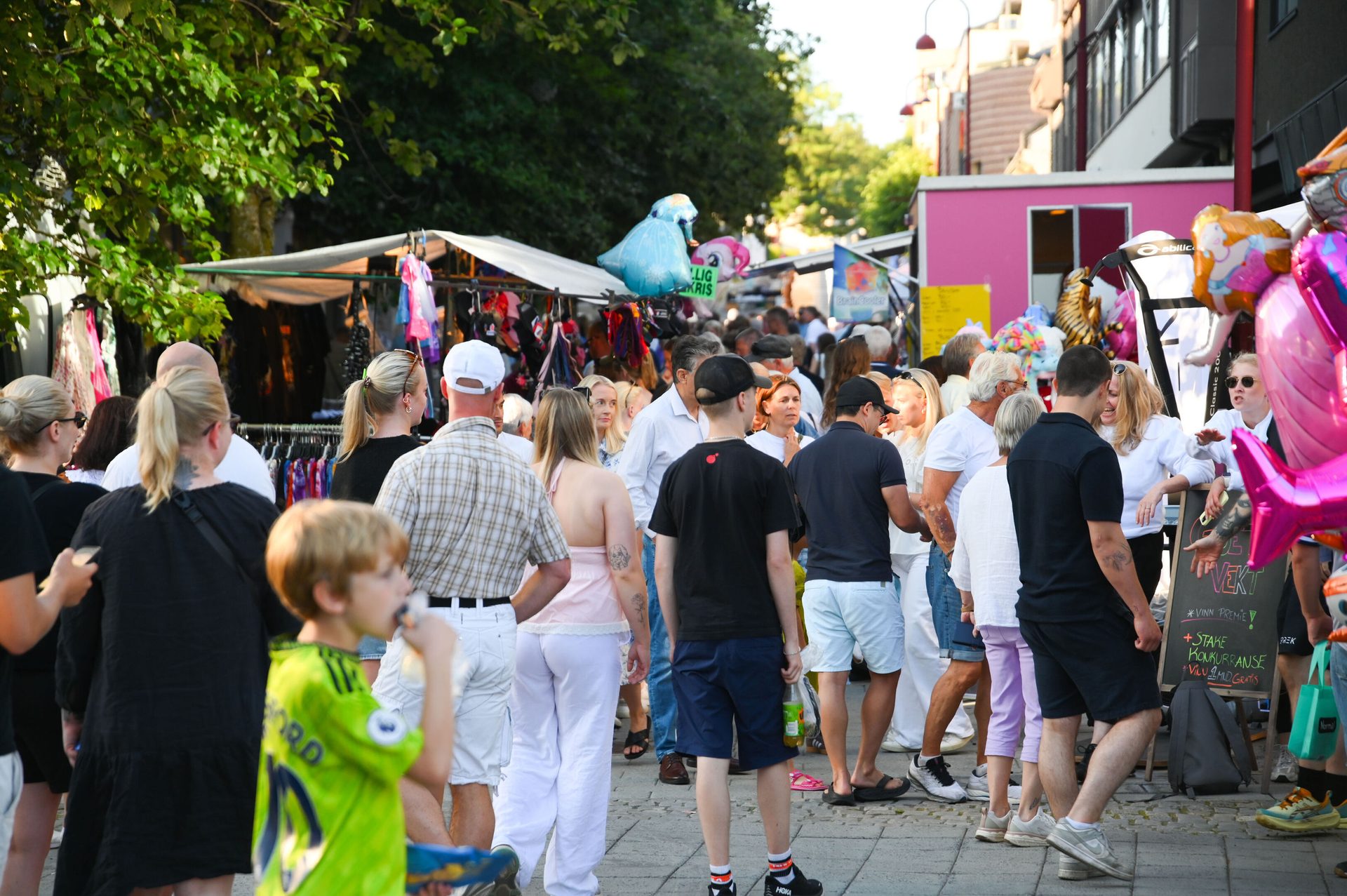 Crowd of people at a bustling outdoor market with colorful stalls and balloons.