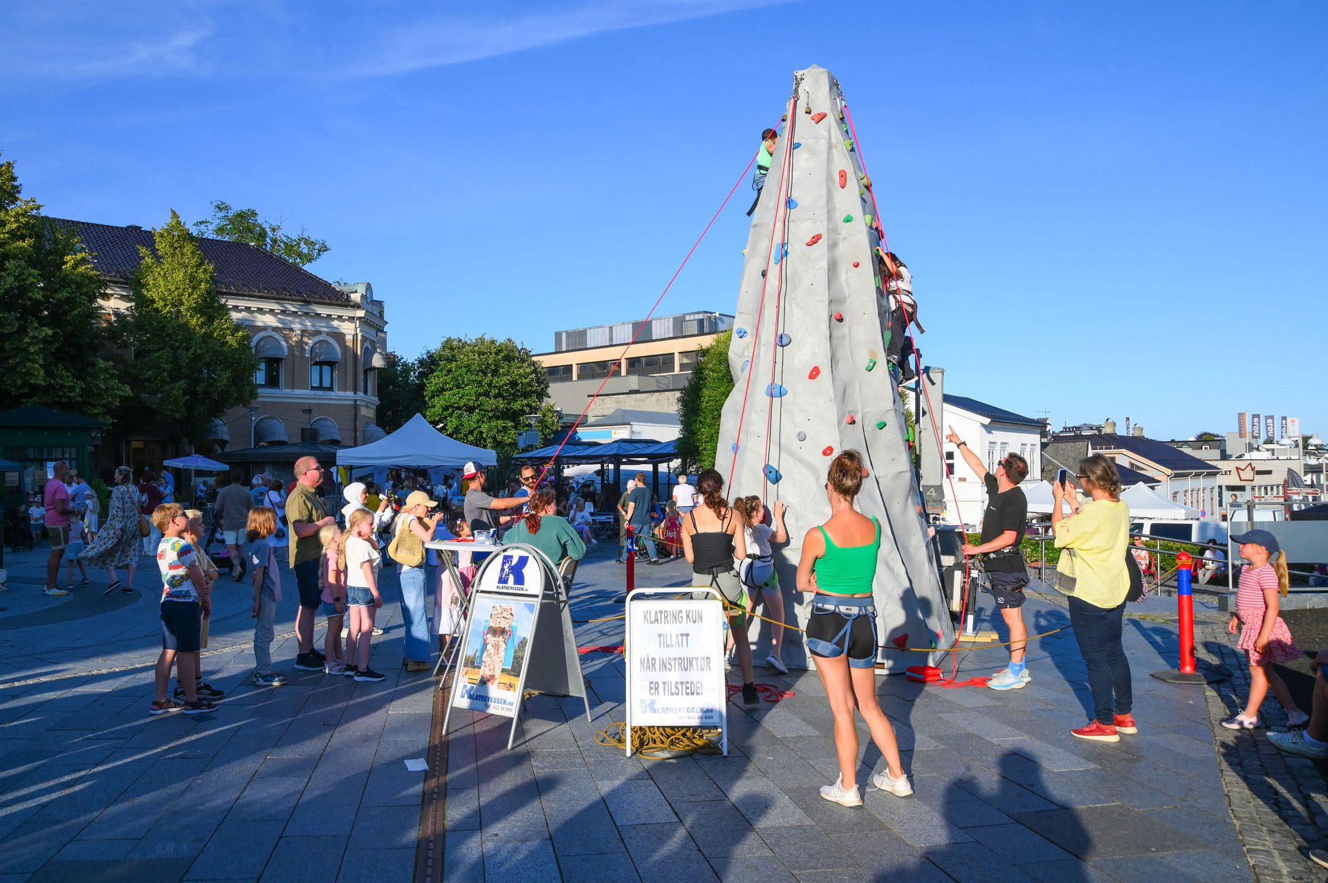 People climb an outdoor rock wall at a lively event on a sunny day.