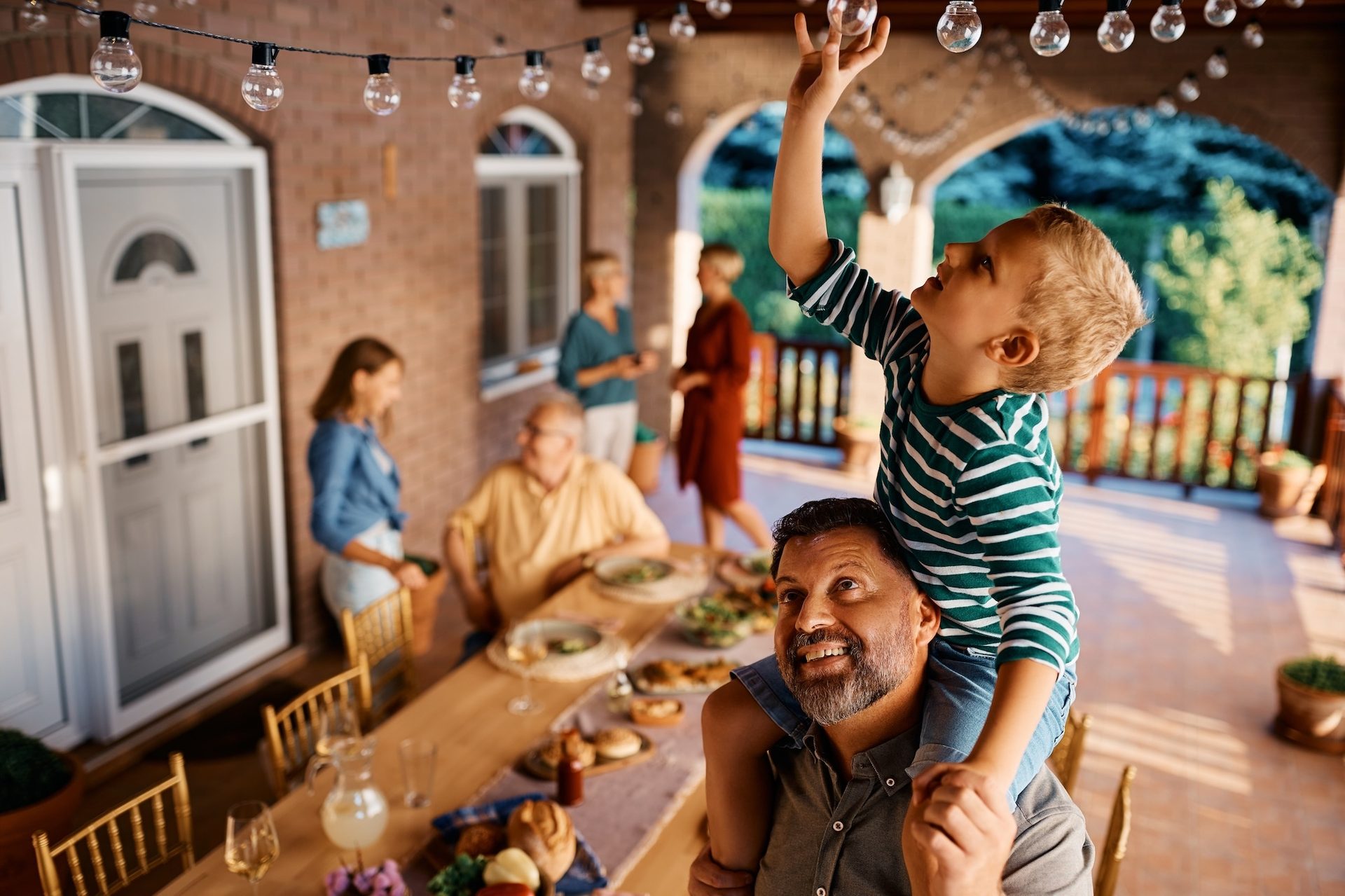 A boy on a man's shoulders reaches for string lights during a family outdoor dinner.