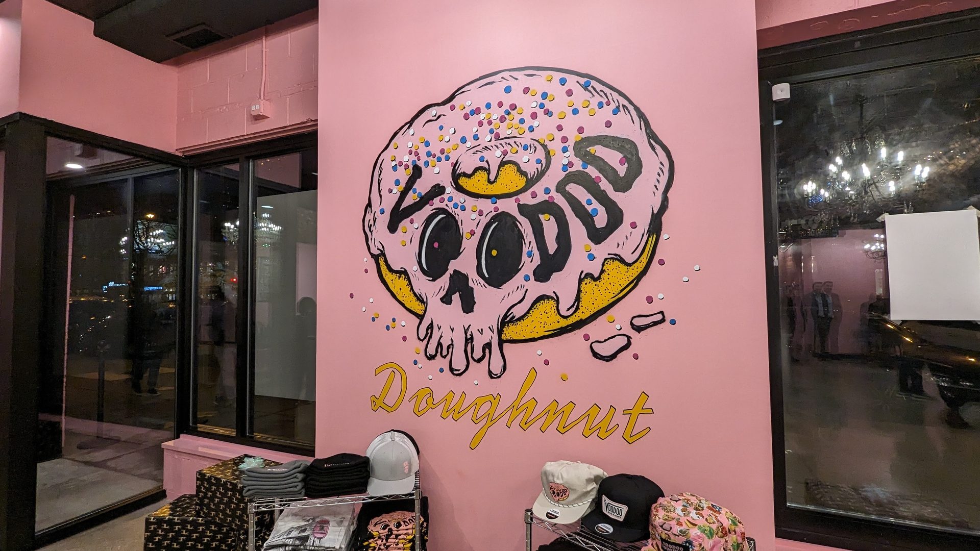 Pink wall with a large Voodoo Doughnut skull mural and merchandise below.