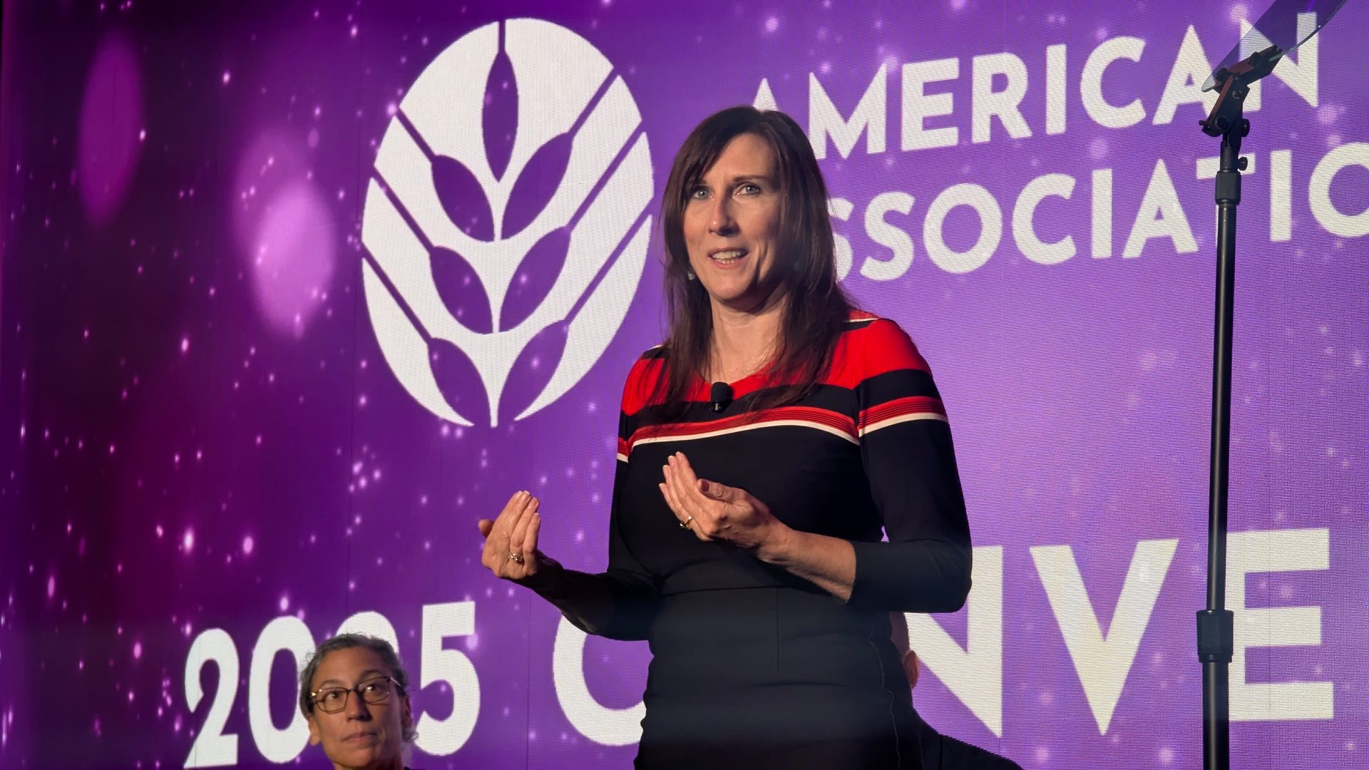 A woman speaks on a stage with a purple screen displaying a logo and "AMERICAN ASSOCIATION" text.