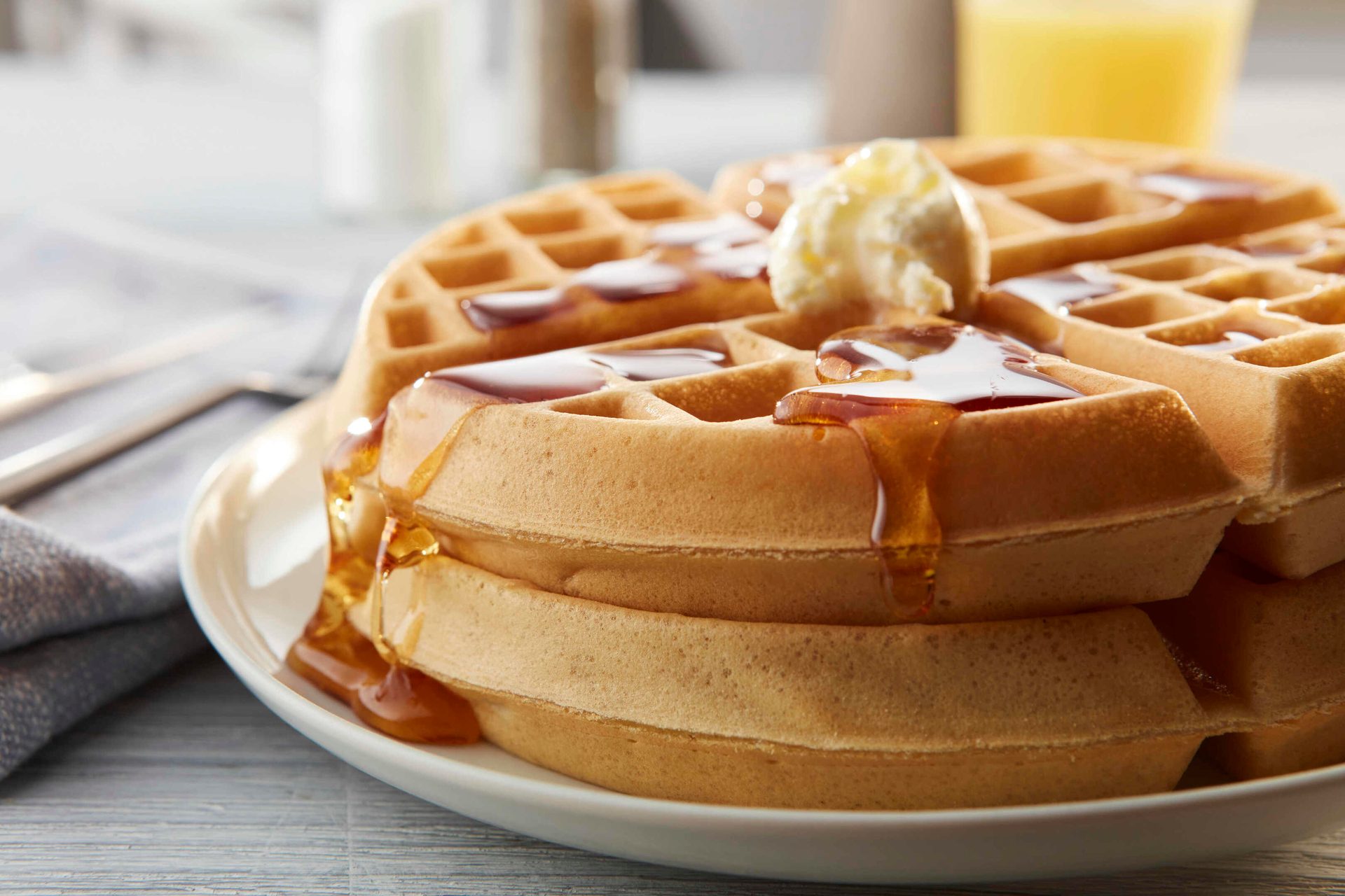 Close-up of golden waffles stacked on a white plate, topped with melting butter and dripping maple syrup.