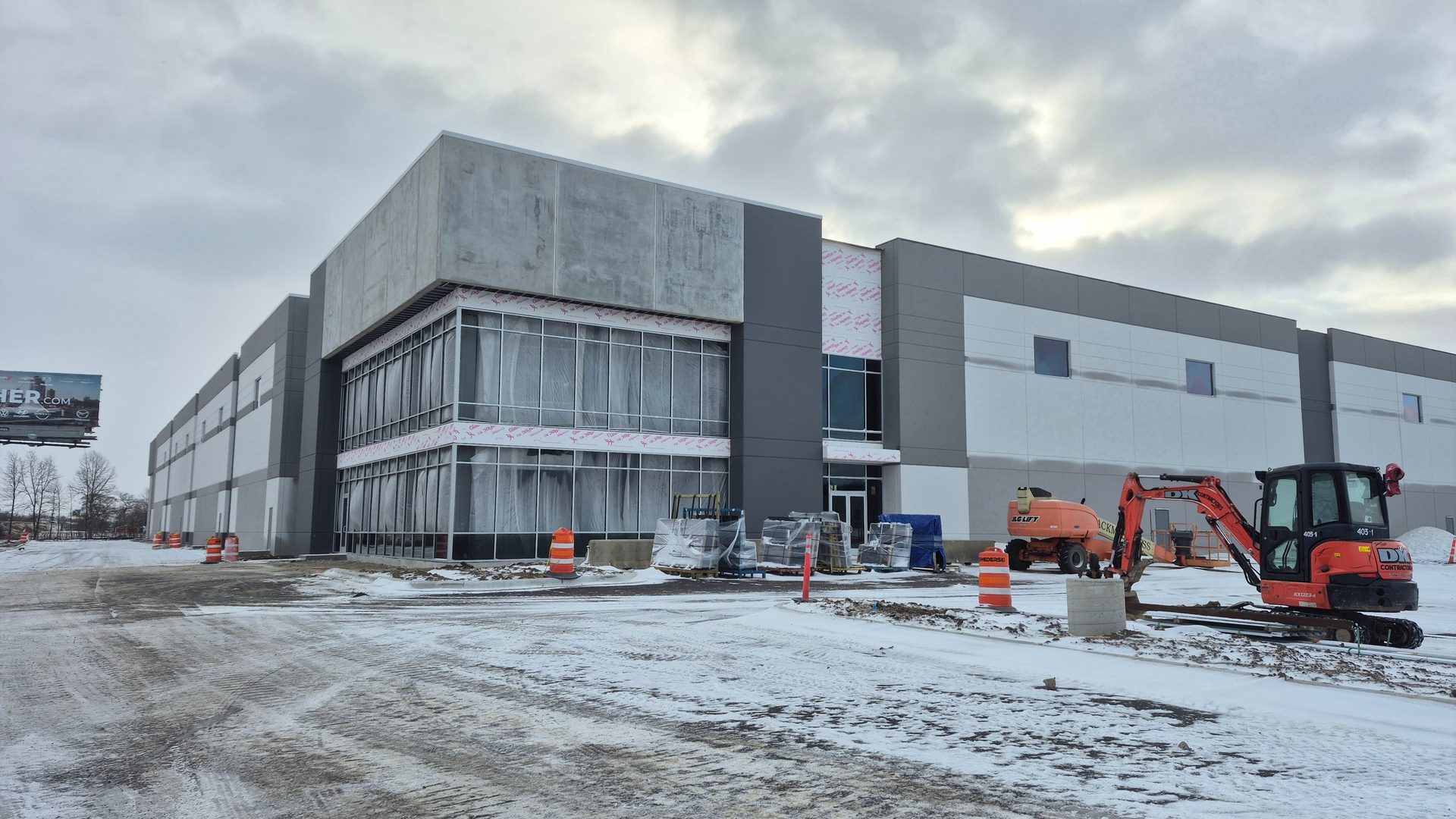 Modern warehouse building under construction on a snowy day with an excavator and a lift.