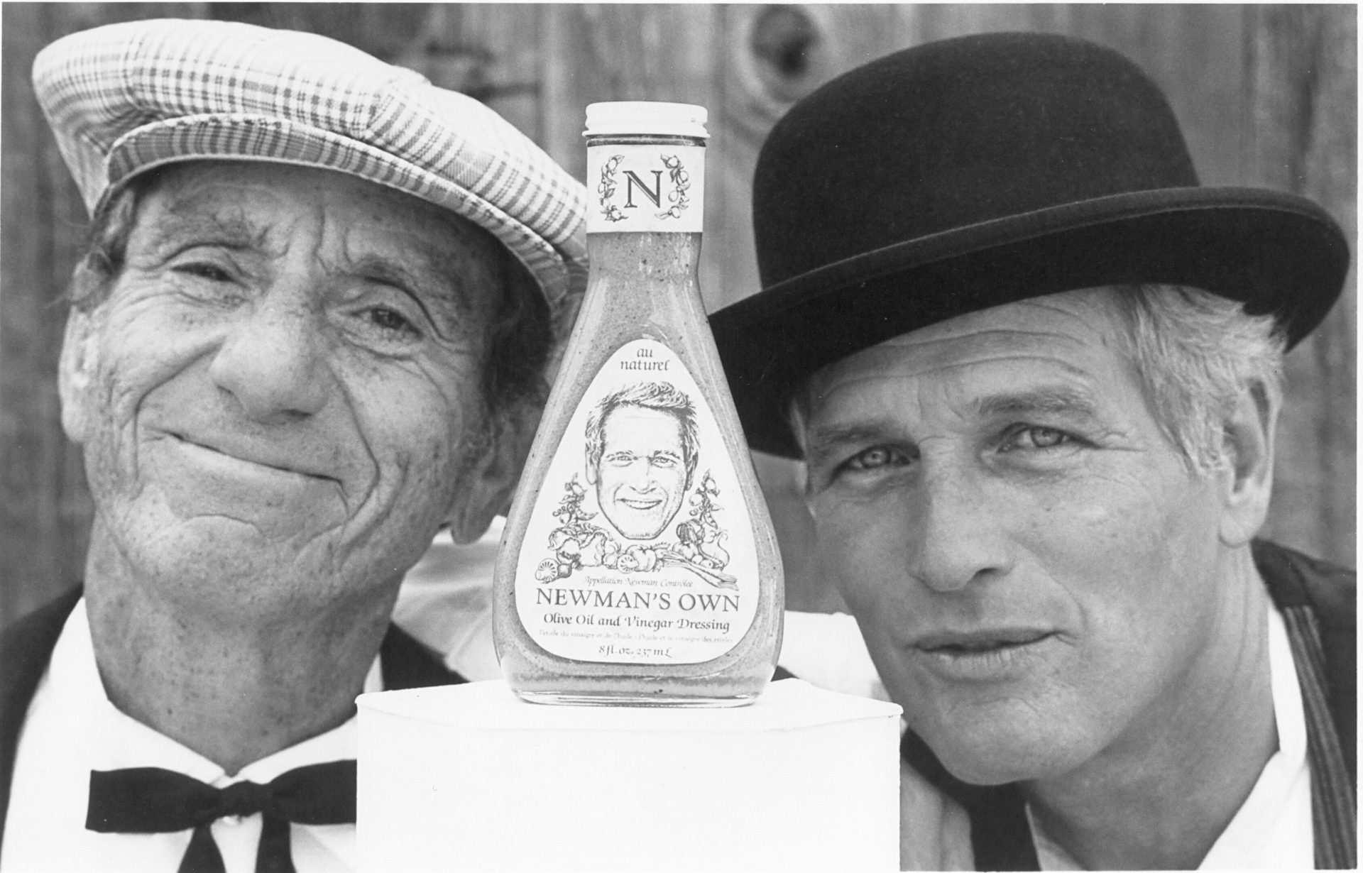 Paul Newman and A. E. Hotchner flank a Newman's Own dressing bottle in a black and white photo.