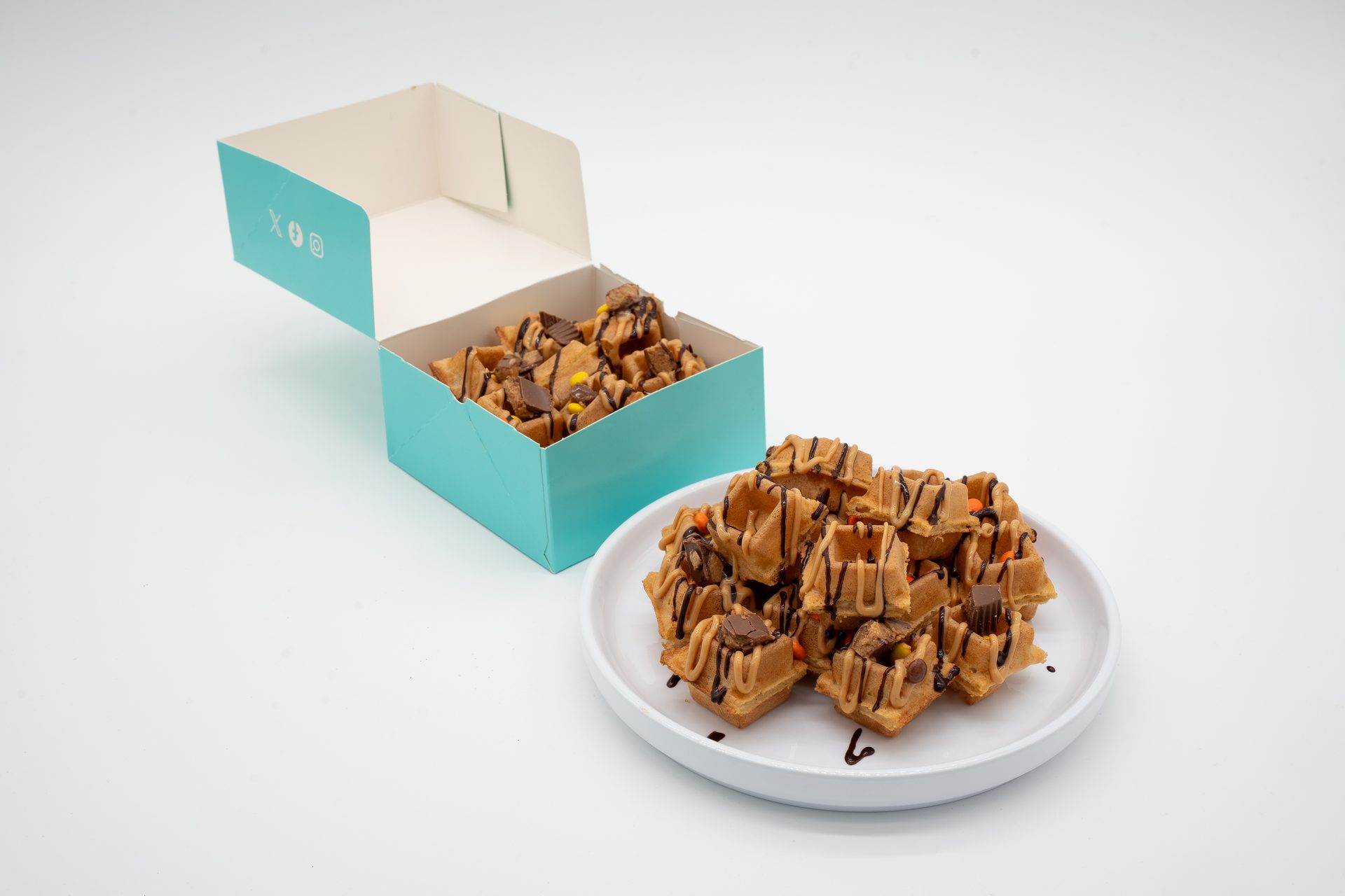 Mini waffle bites with chocolate drizzle and peanut butter candy, some in a teal box, some on a white plate.