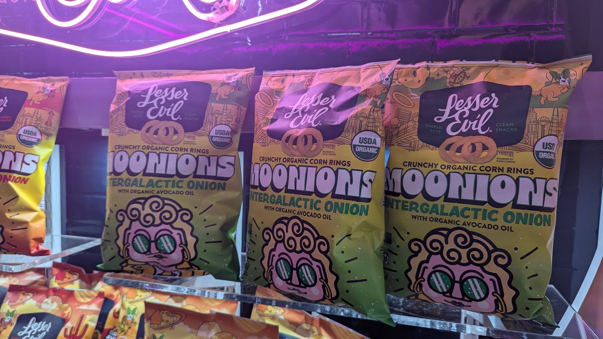 Bags of Lesser Evil Moonions Intergalactic Onion organic corn rings under purple neon light.