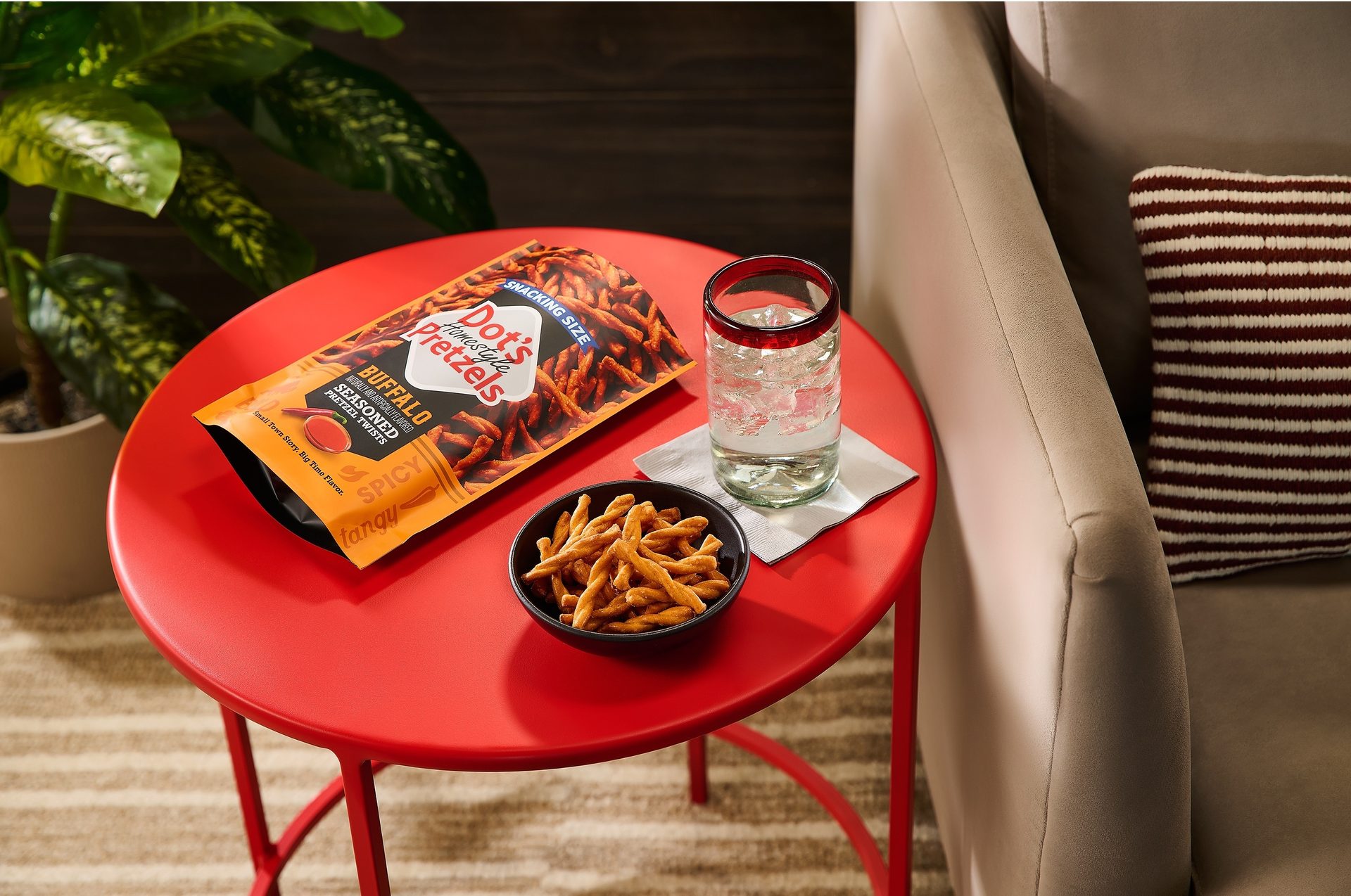 A bag of Dot's Buffalo Seasoned Pretzels, a bowl, and a glass of water on a red side table.