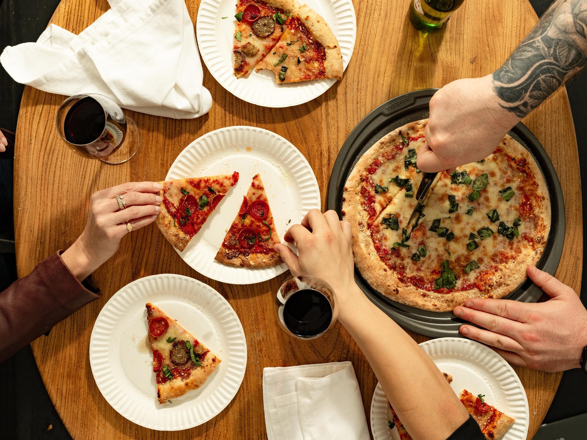 Overhead view of multiple people sharing and eating different kinds of pizza slices on a wooden table.