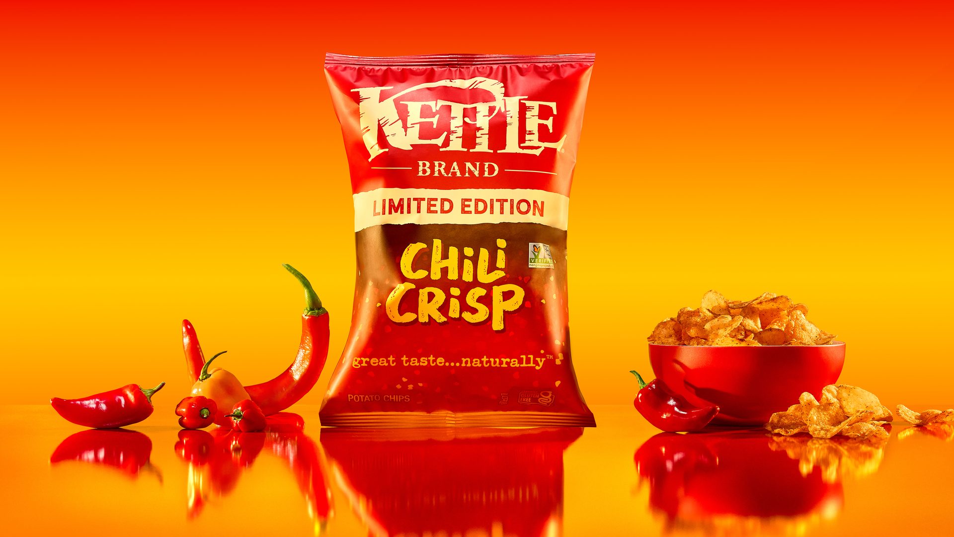 Kettle Brand Chili Crisp chips bag, red chili peppers, and a bowl of chips on a fiery red-orange surface.