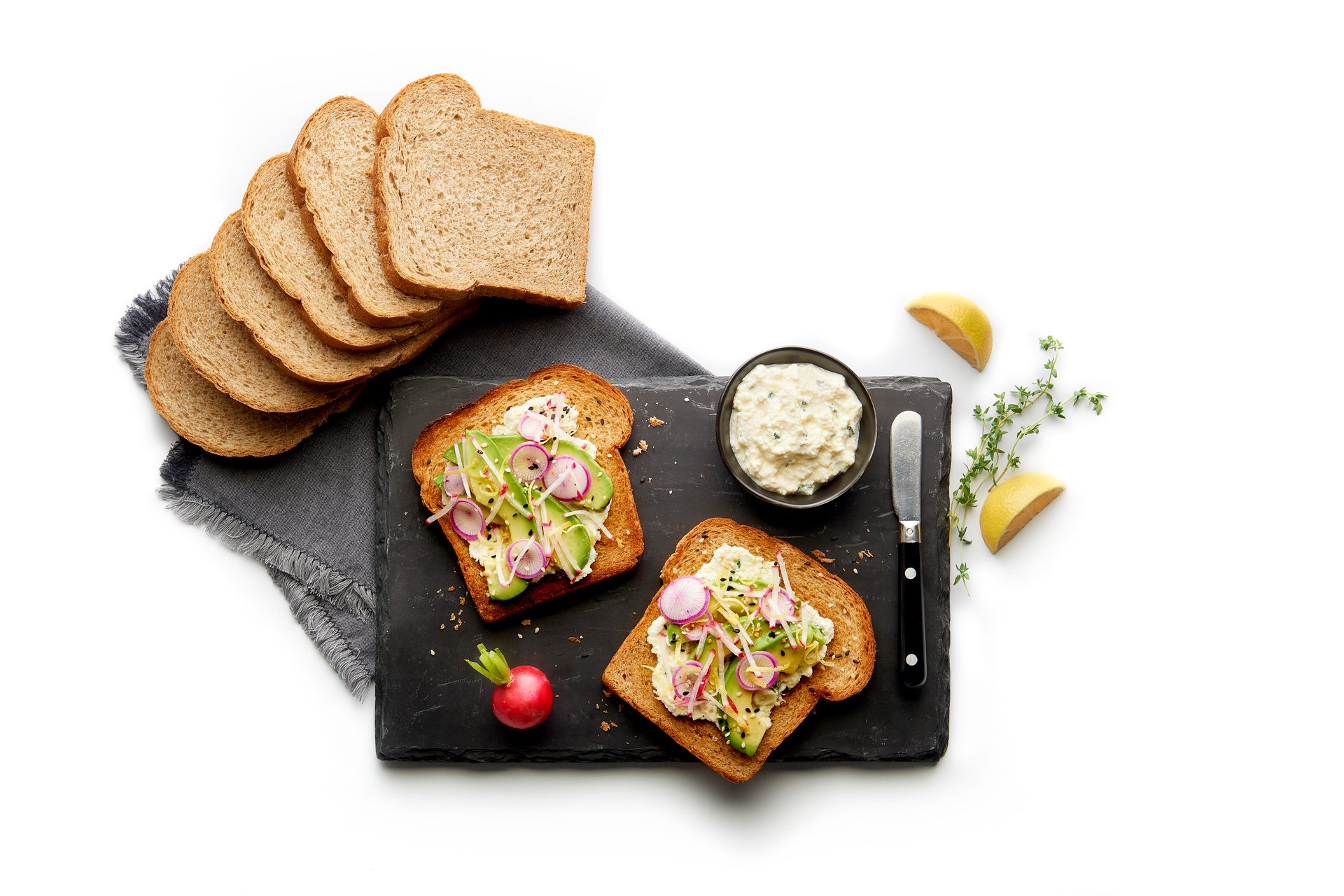 Toasted bread topped with avocado, radishes, and spread, with more bread slices, lemon, and herbs.