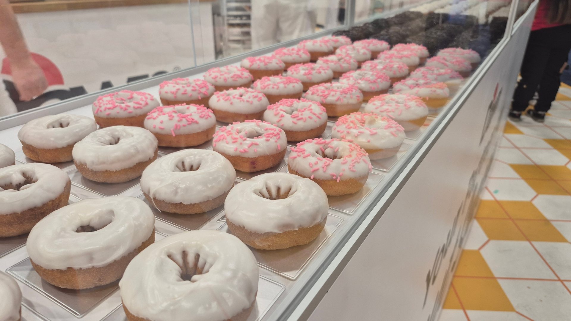 Rows of white-iced donuts, some with pink sprinkles, displayed in a glass case.