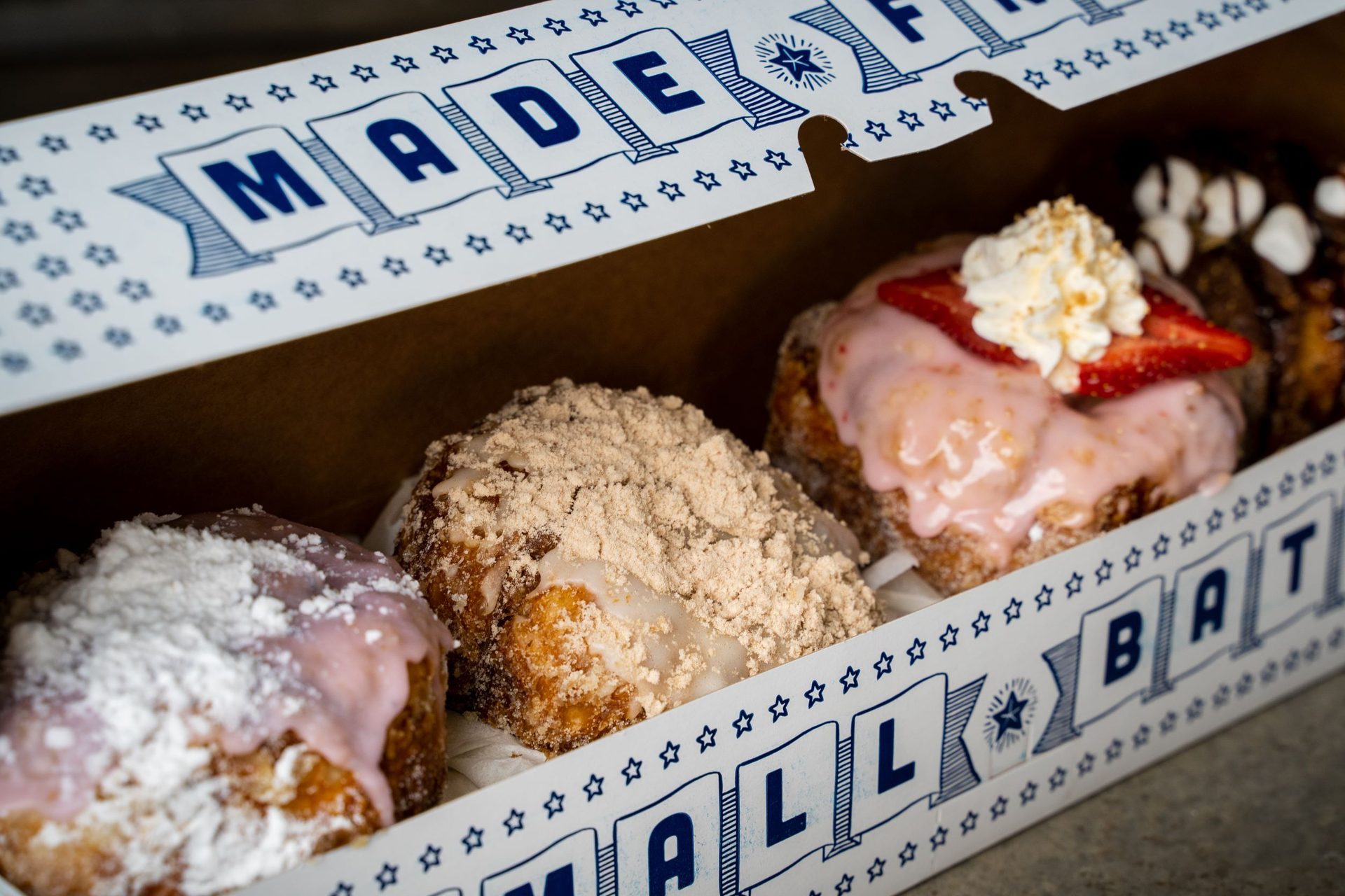 A box of assorted gourmet donuts with various toppings like strawberry and crumble, featuring brand text.