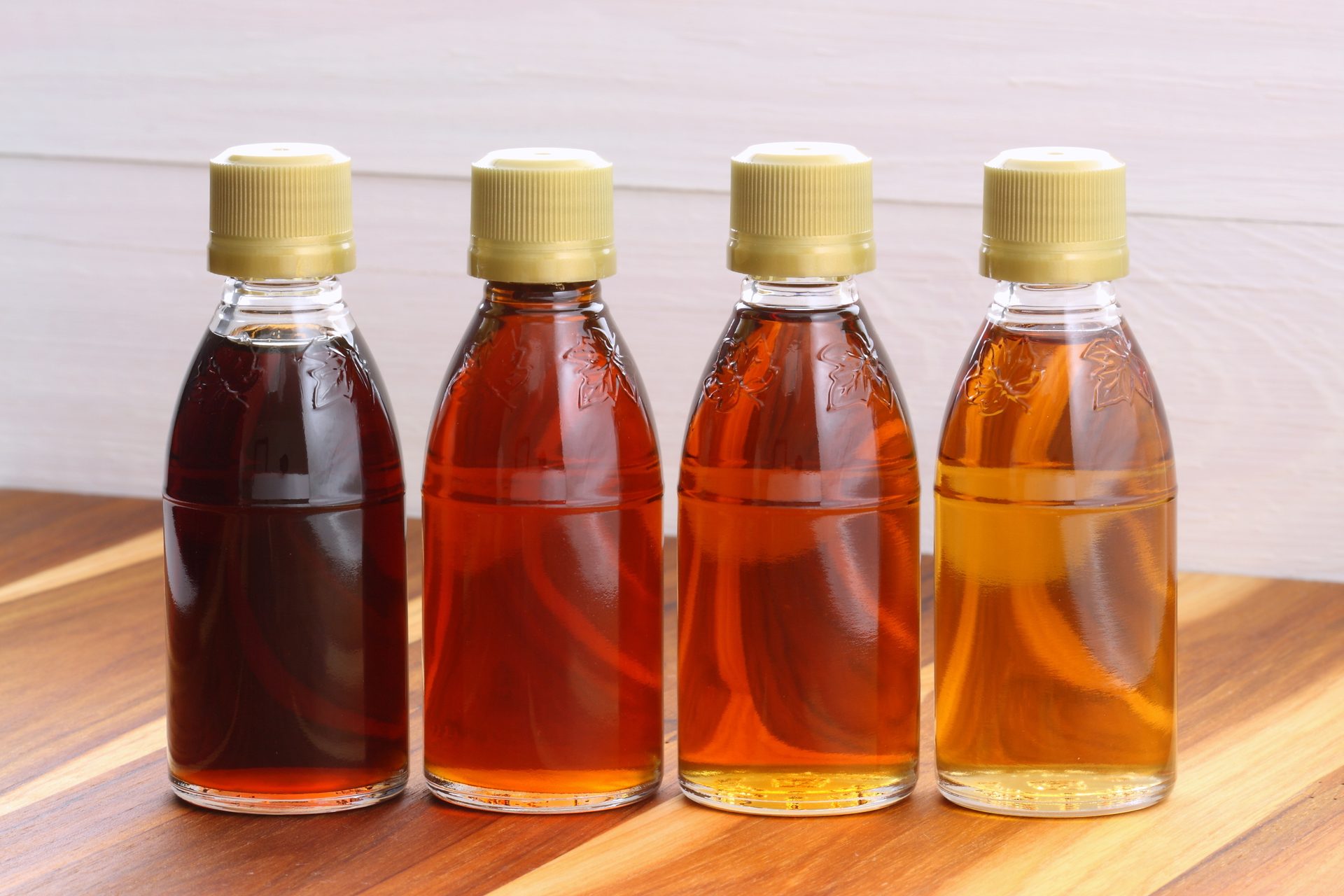 Four bottles of maple syrup, ranging from dark to light amber, on a wooden surface.