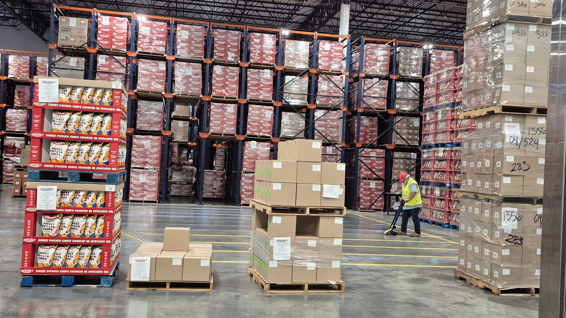 Large warehouse with pallets of chips and boxes, a worker pushing equipment, and tall storage racks.