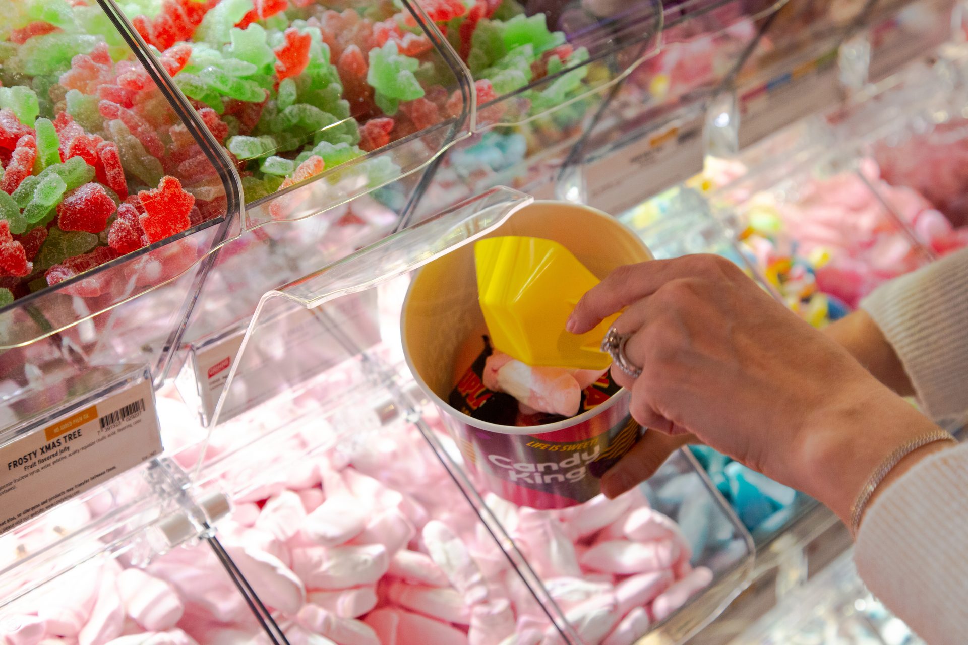 Hand fills a 'Candy King' cup with red and green gummy candies and pink marshmallows from bulk bins.
