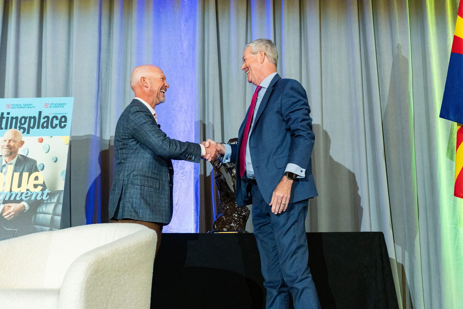 Two smiling men in suits shake hands on a stage.