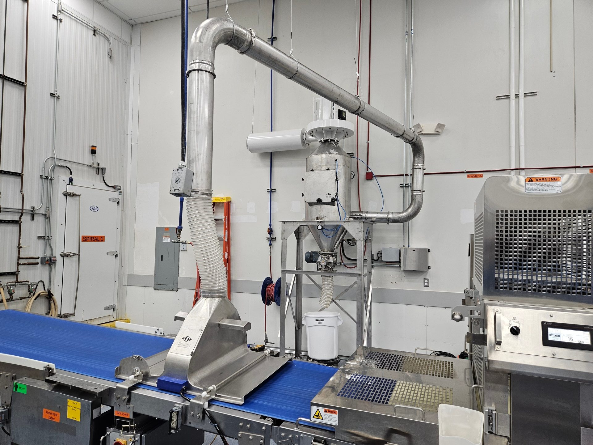 Industrial food processing room with a blue conveyor belt, stainless steel machinery, and overhead ducts.