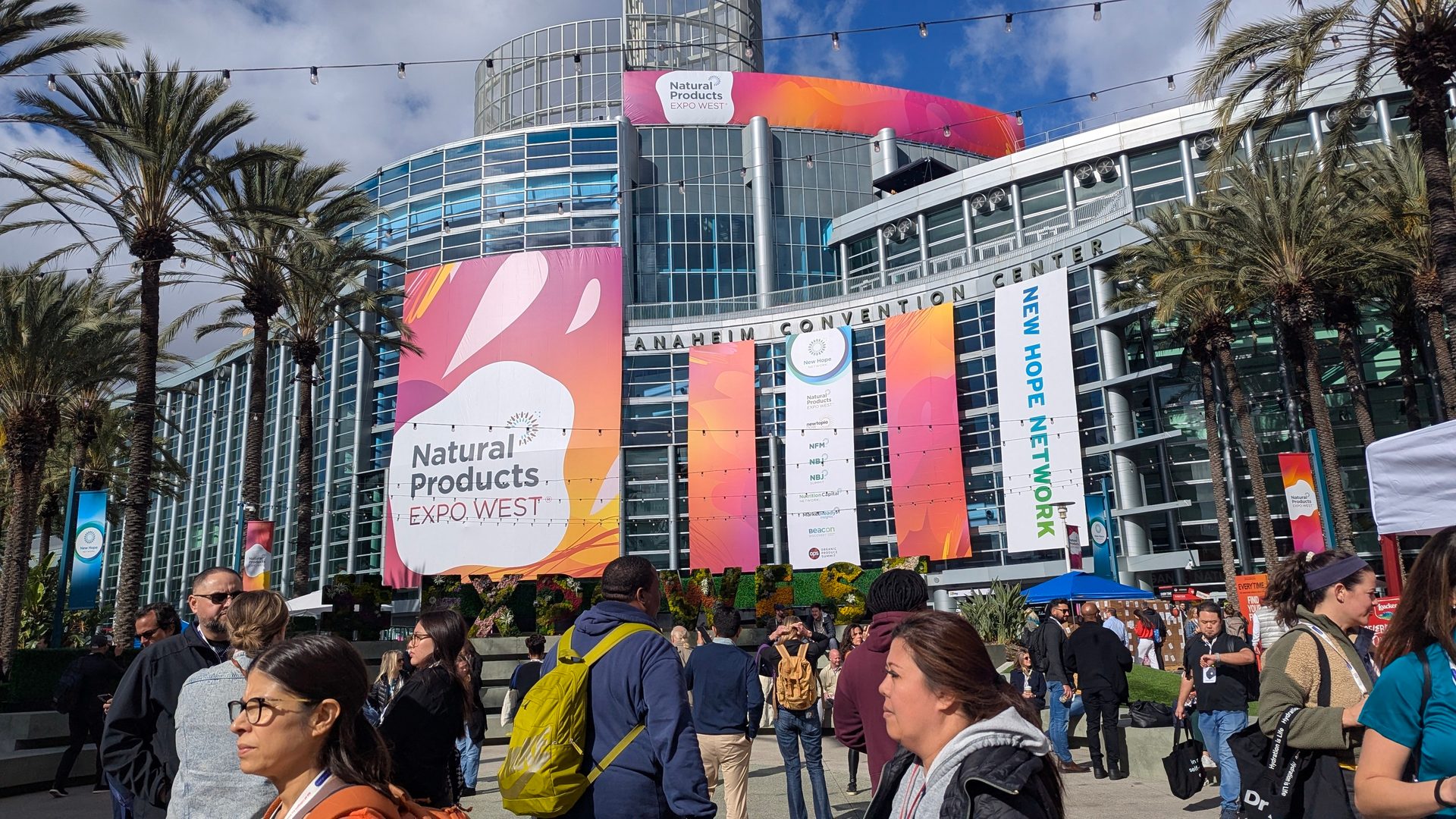 Crowd outside Anaheim Convention Center with "Natural Products Expo West" banners and palm trees.