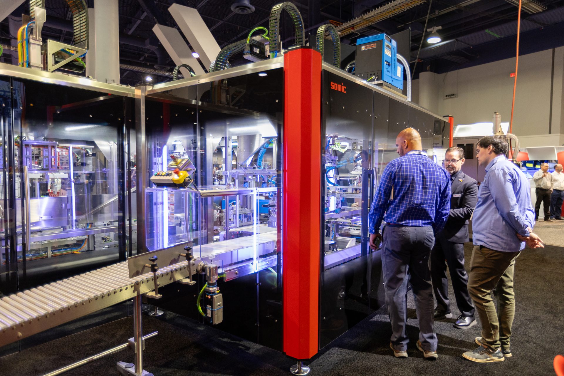Three men observe a large SOMIC industrial packaging machine at an exhibition booth.