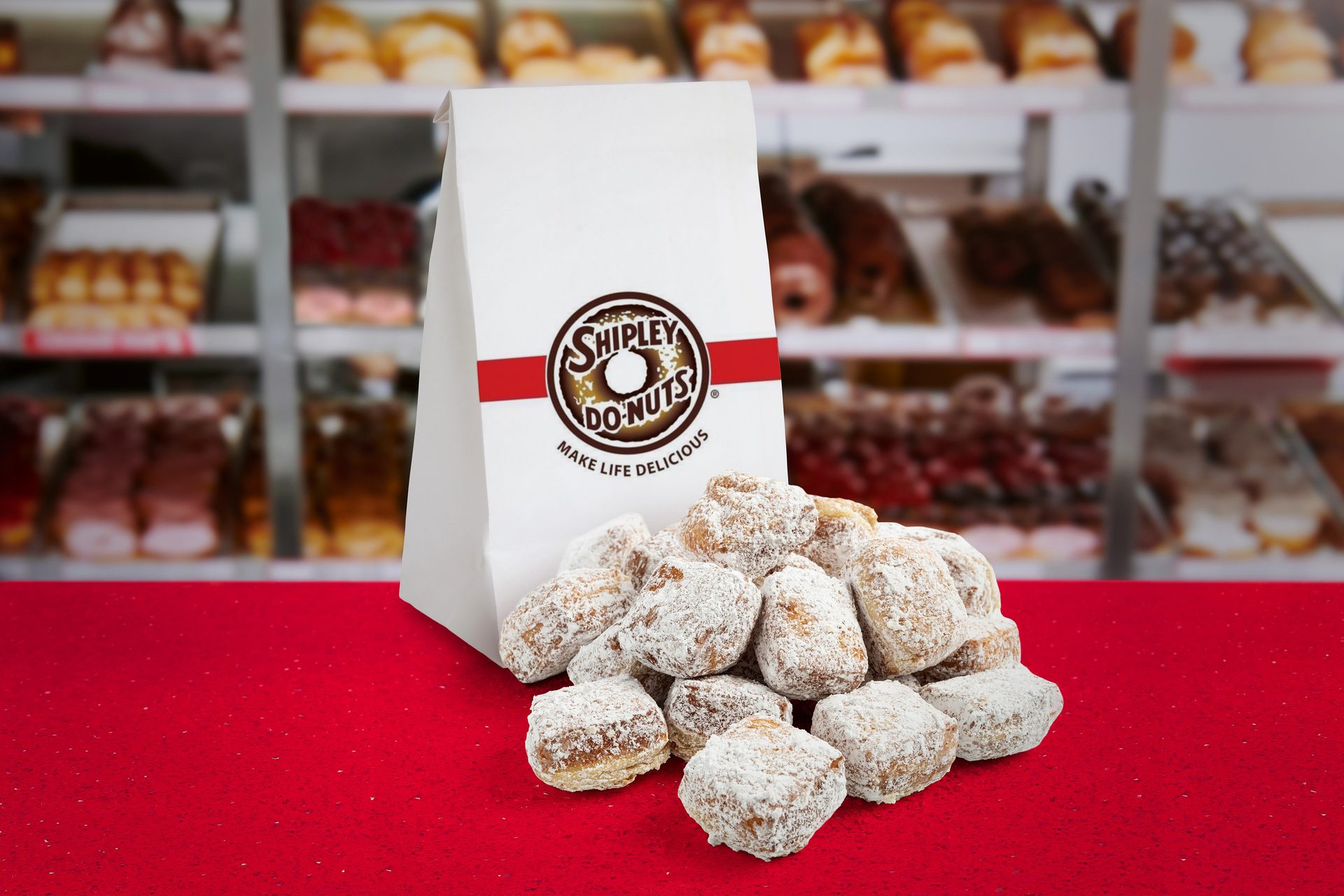 White Shipley Do-Nuts bag with a pile of powdered donut holes on a red table, donut display blurred in background.