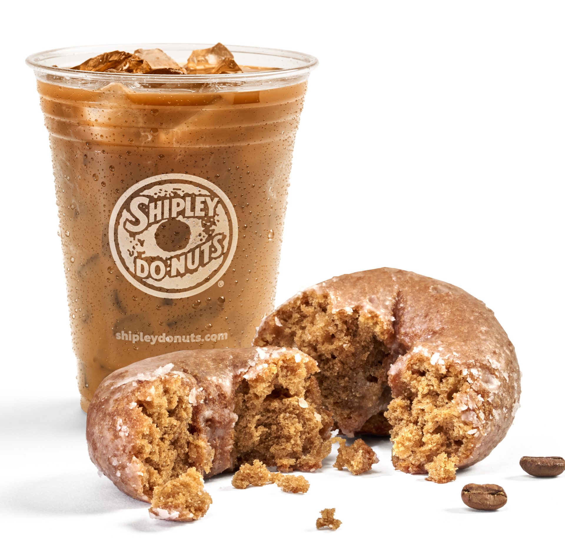 Iced coffee in a Shipley Do-Nuts cup, broken donut, and coffee beans on white.