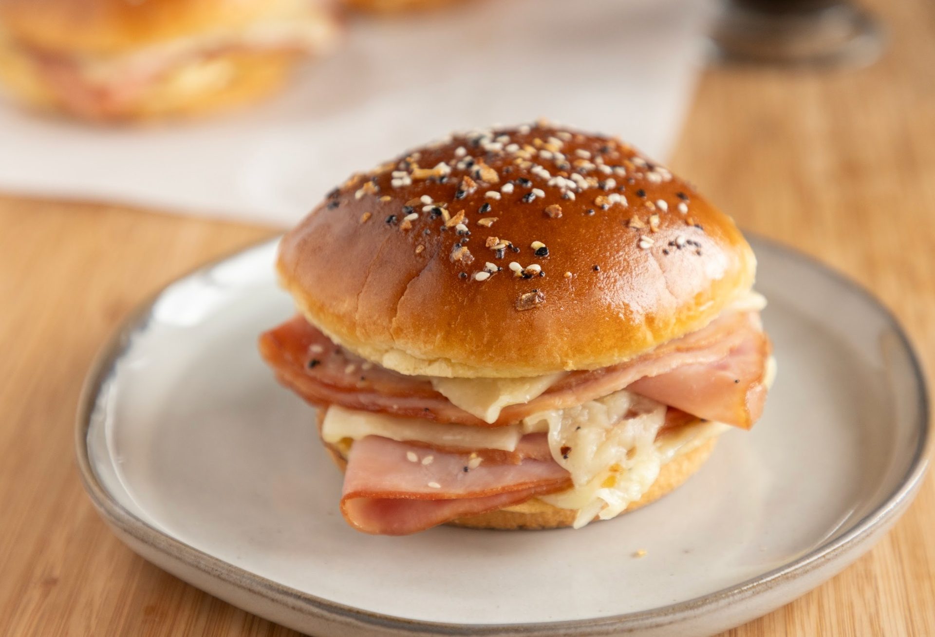 A ham and melted cheese slider on an everything bagel seasoned bun, on a grey plate.