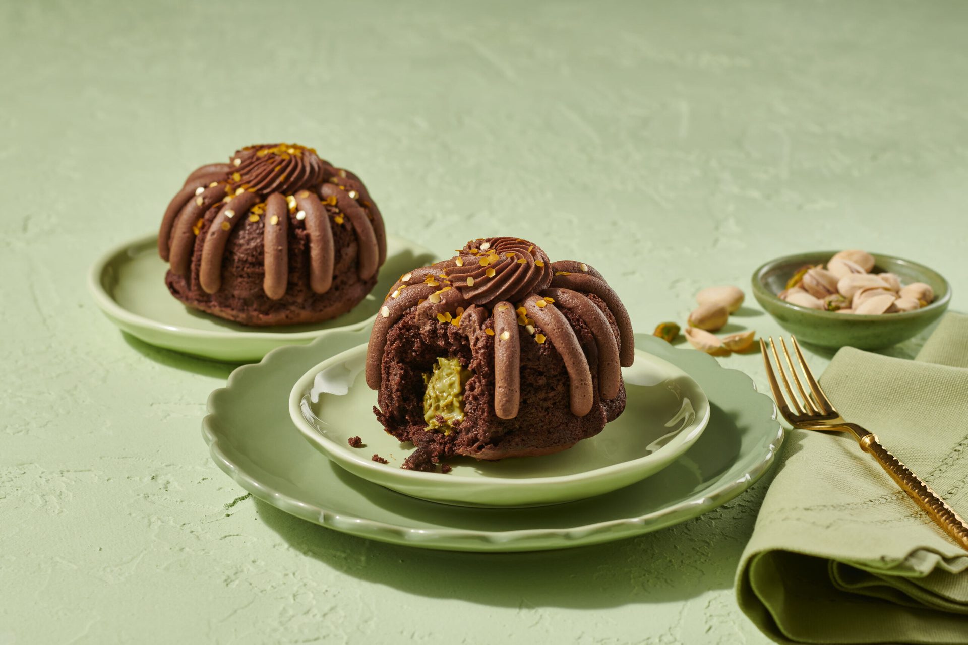 Two chocolate bundt cakes with pistachio filling, served on green plates with pistachios and a gold fork.