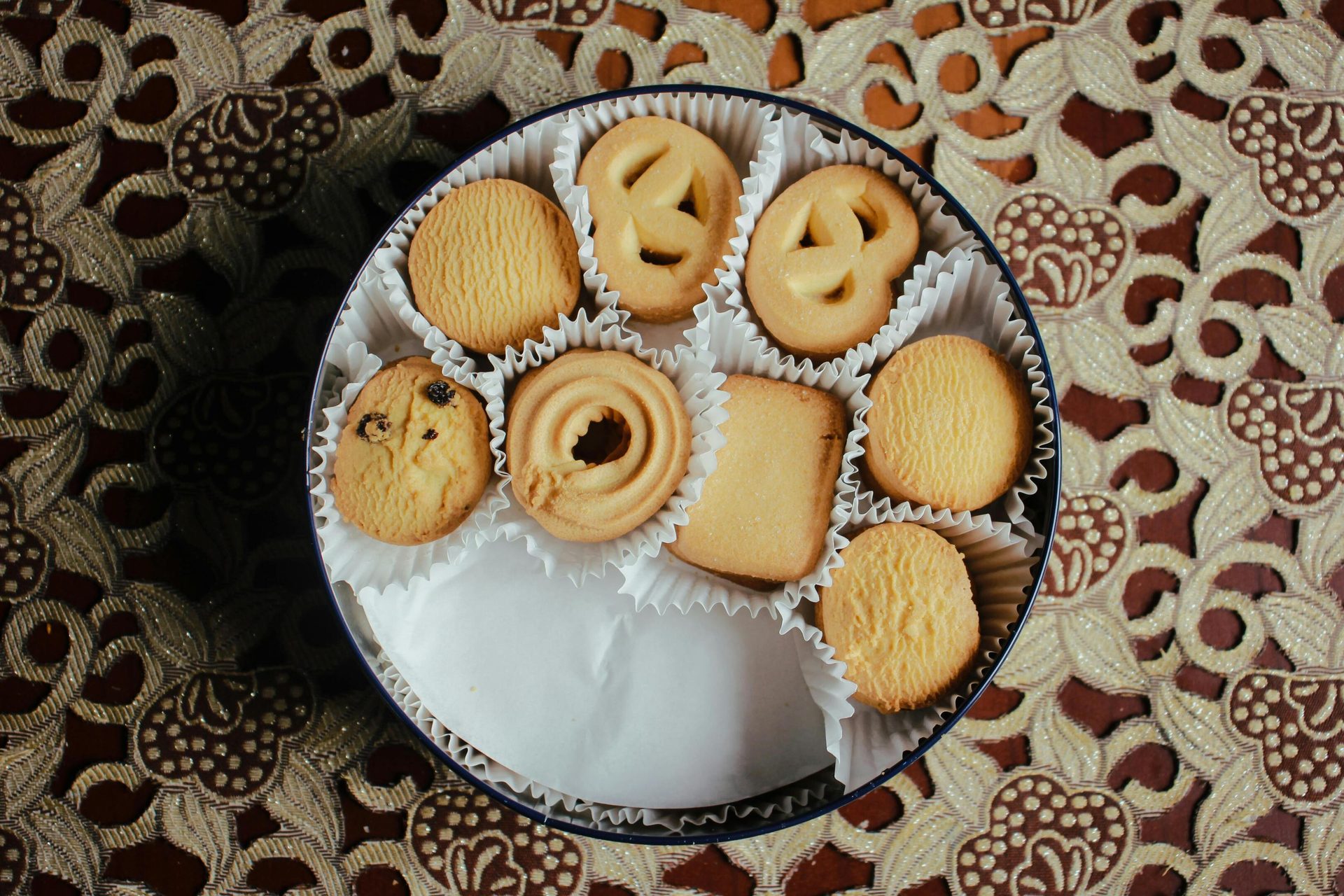 A round tin of assorted butter cookies on a brown and gold patterned tablecloth.