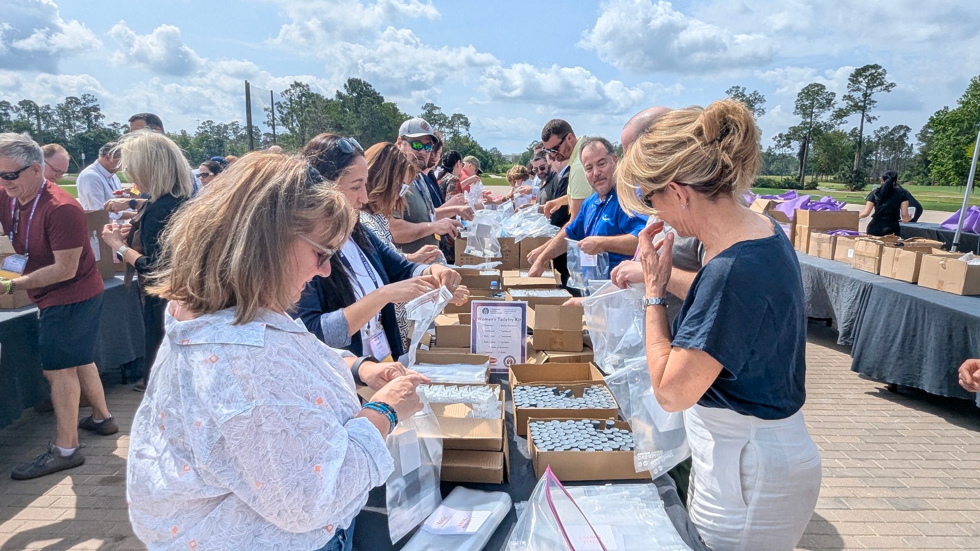People pack toiletry kits into bags at a long outdoor table under a cloudy sky, with trees in the background.