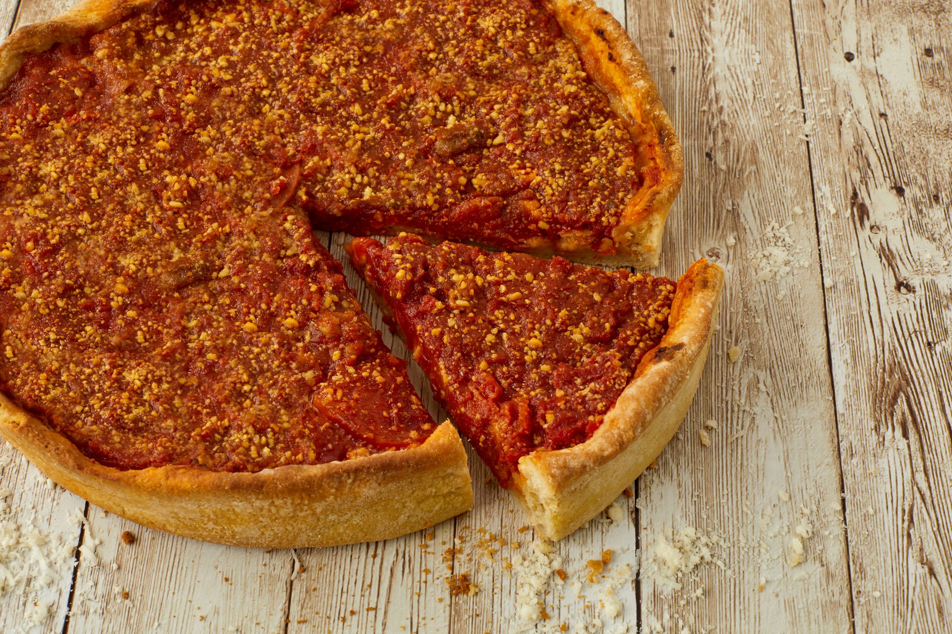 Deep-dish pizza with a rich red sauce and crumbled topping, with one slice removed, on a wooden table.