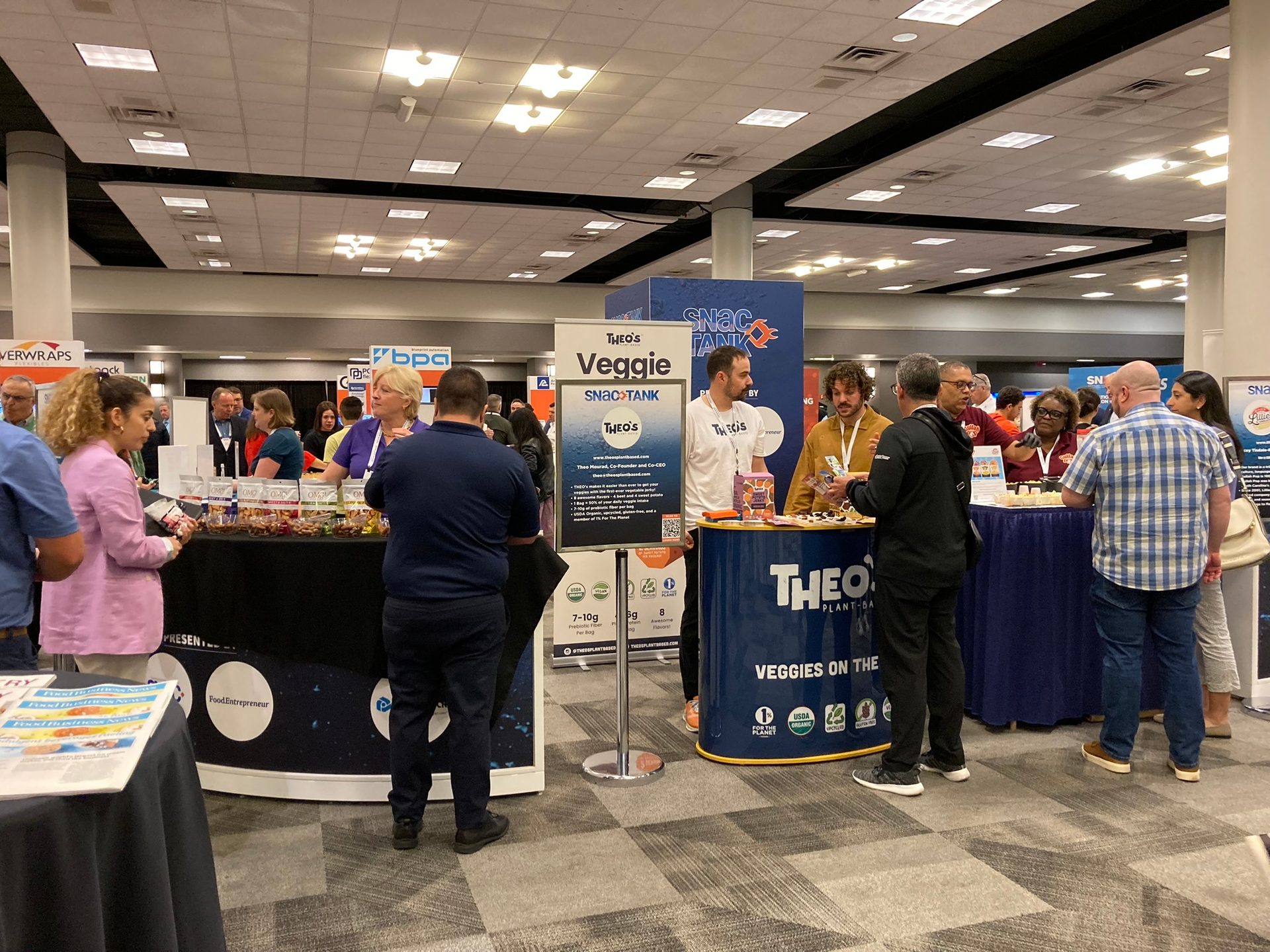 People interacting at booths at an indoor food trade show, with a prominent "Theo's Veggie SNAC-TANK" display.