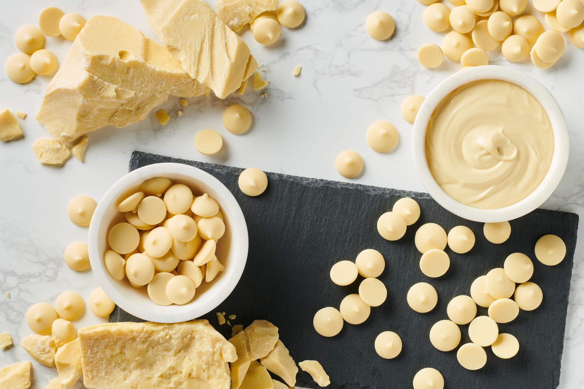 Cocoa butter chunks, white chocolate chips, and melted white chocolate in bowls on a marble surface.