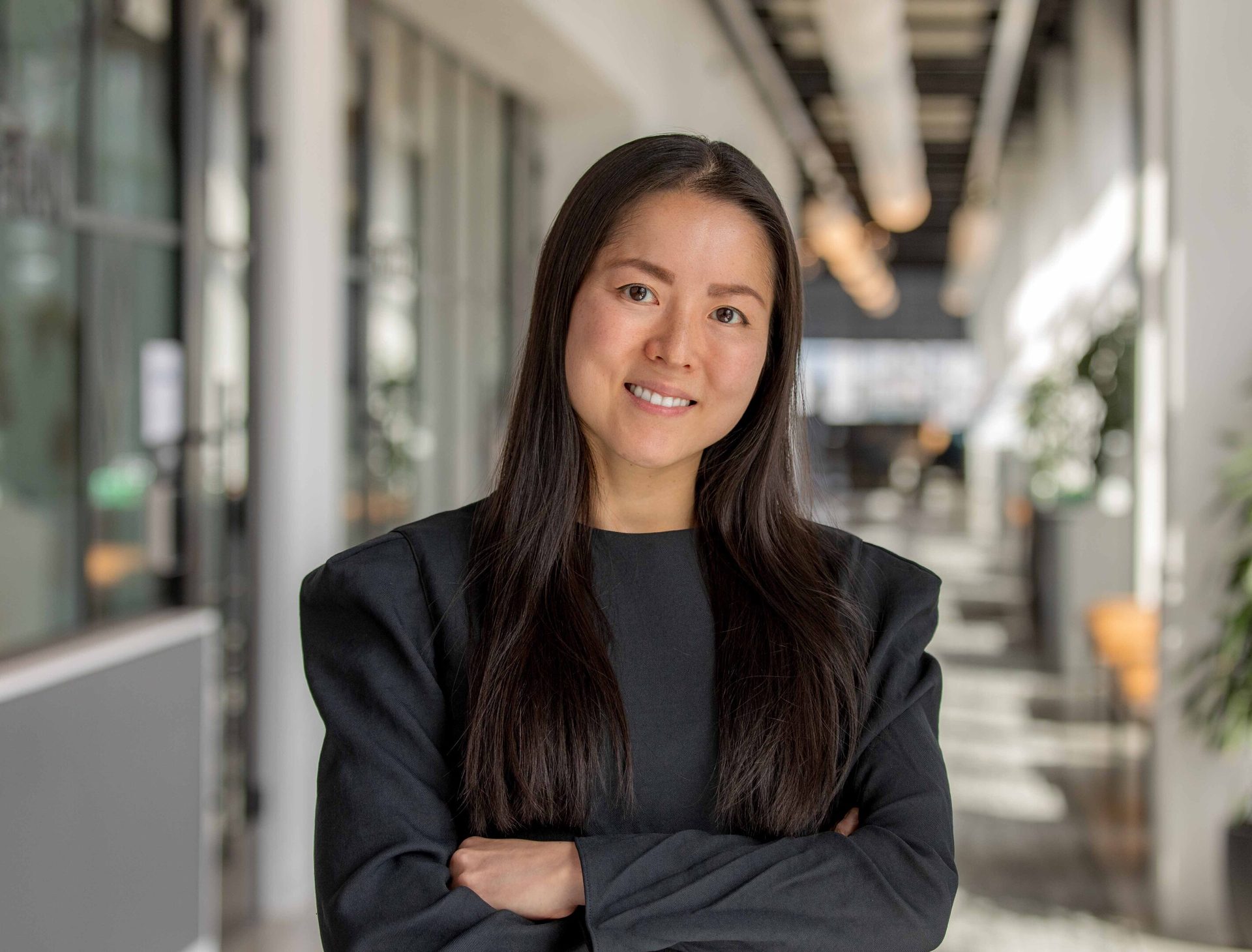 Smiling woman with dark hair, dark top, arms crossed, in a blurred office.