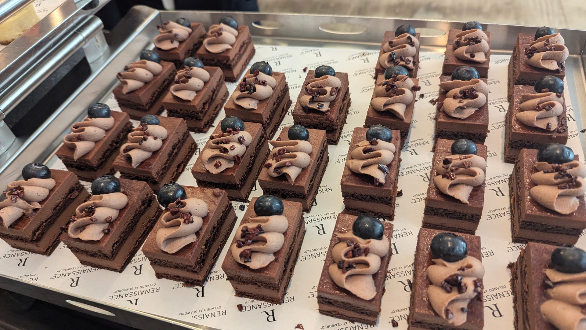 A tray of rectangular chocolate cakes topped with piped chocolate cream, a blueberry, and cacao nibs.