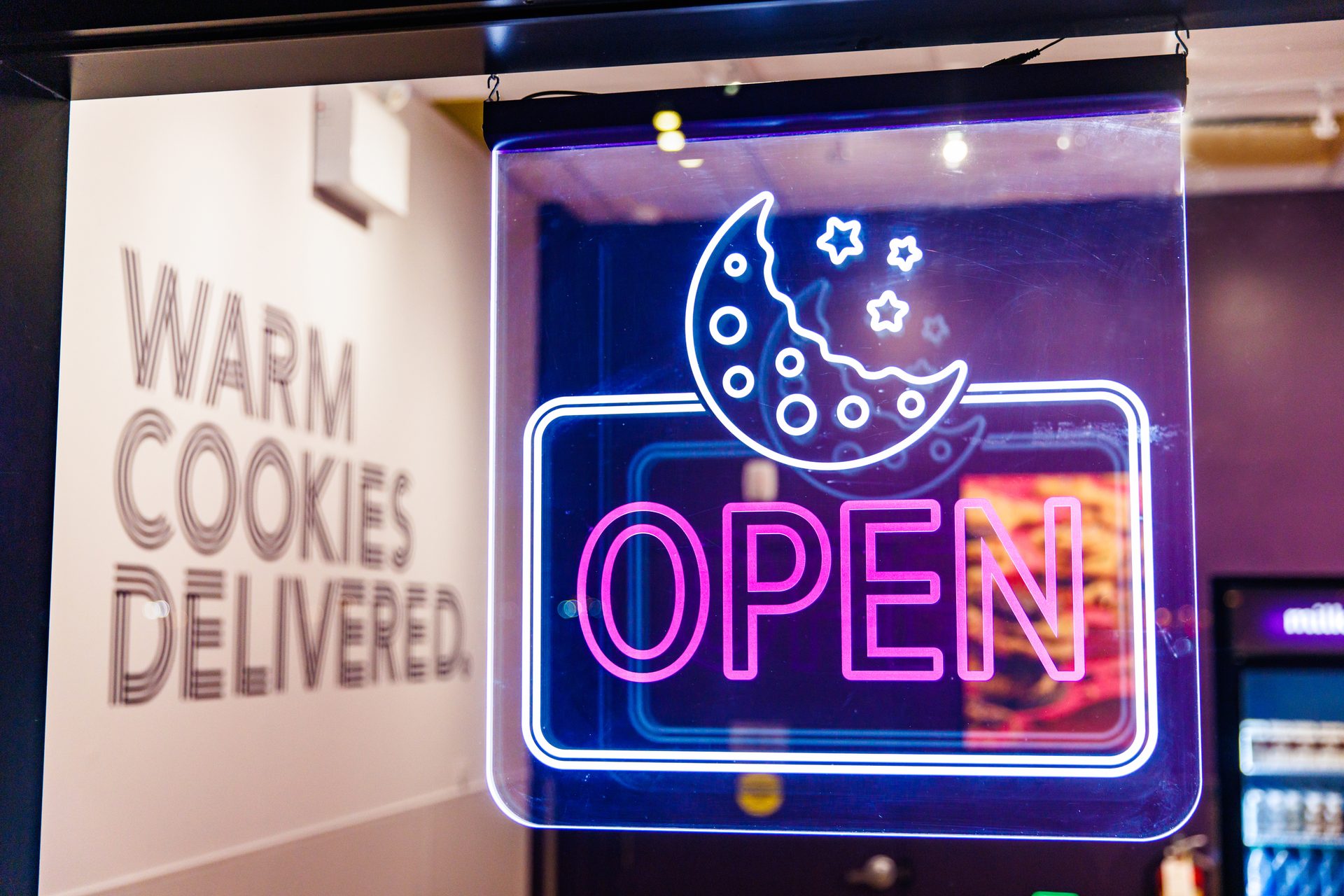 Neon "OPEN" sign with a cookie moon and stars, next to "Warm Cookies Delivered" text on a wall.