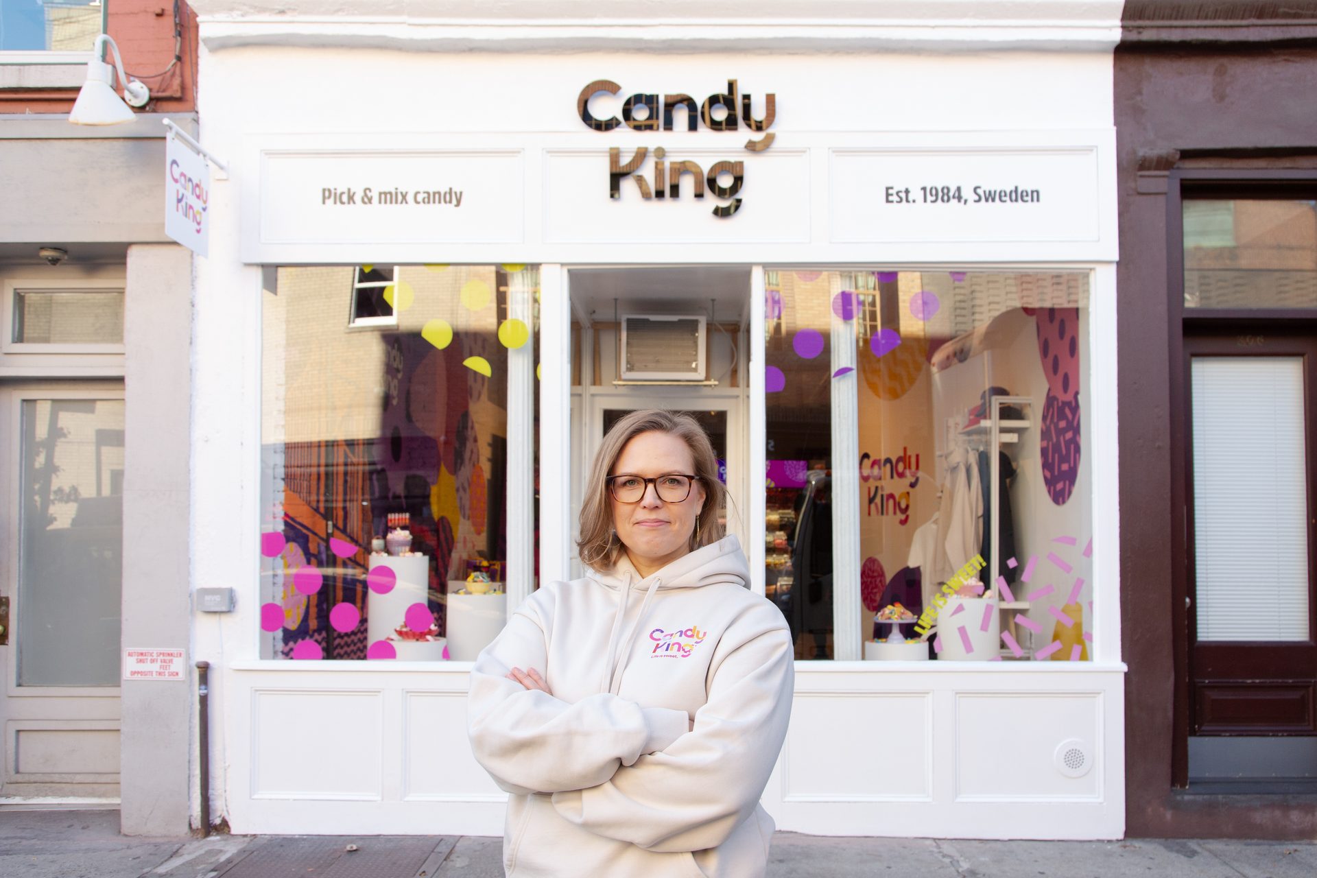 A woman in a "Candy King" hoodie stands with crossed arms in front of the "Candy King" store.