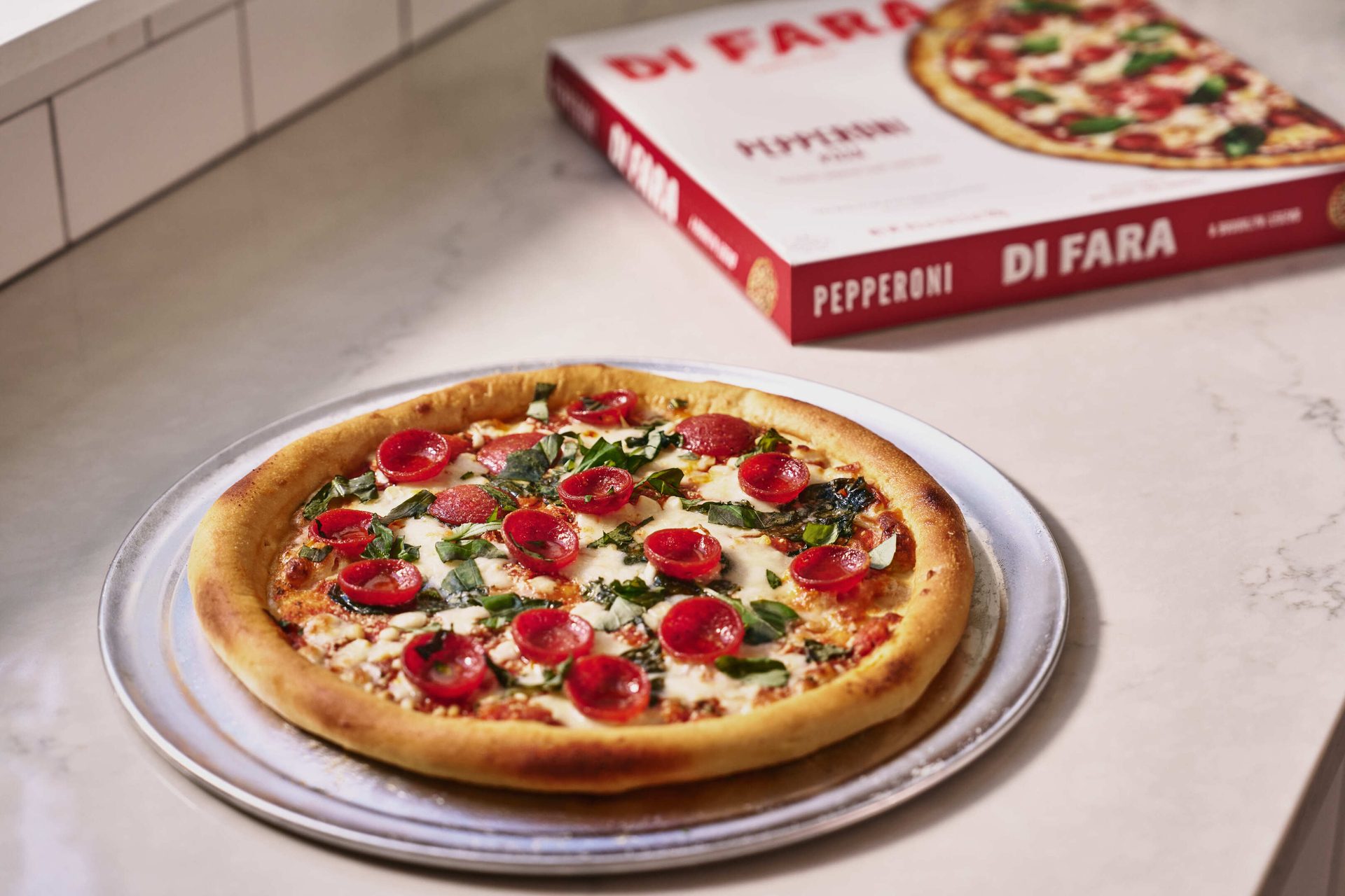A pepperoni pizza with fresh basil on a metal tray, with a Di Fara frozen pizza box in the background.