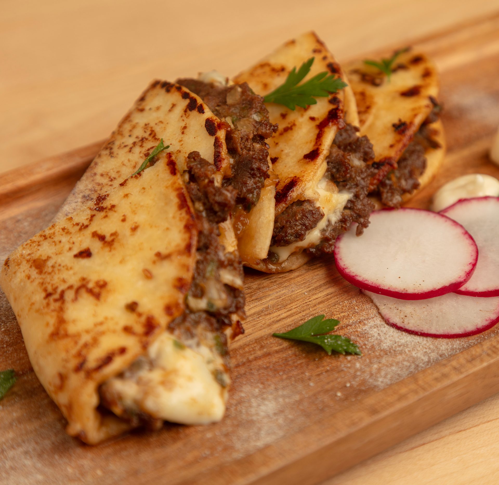 Three cheesy beef quesabirrias with charred tortillas, garnished with parsley and sliced radishes on a wooden board.