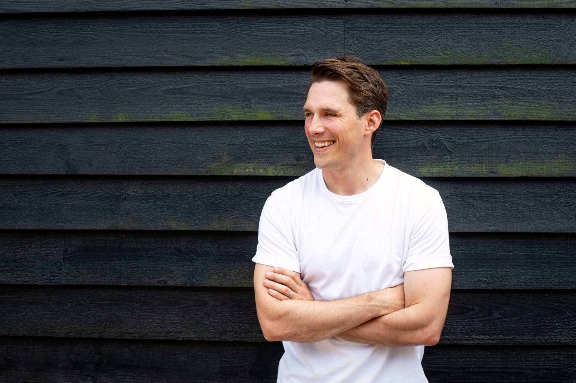 A smiling man in a white t-shirt with crossed arms looks left against a dark wooden wall.