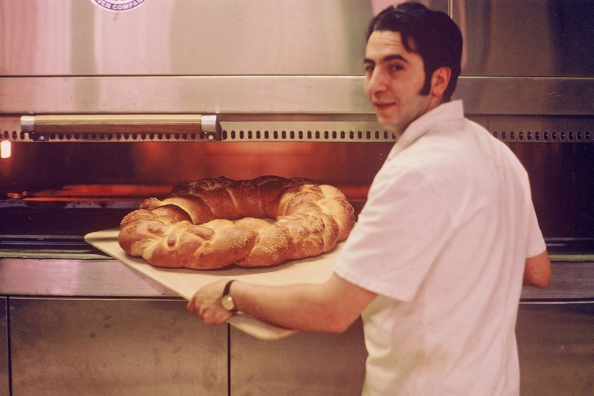 A baker places a large, braided bread into a hot oven with a peel, glancing at the camera.