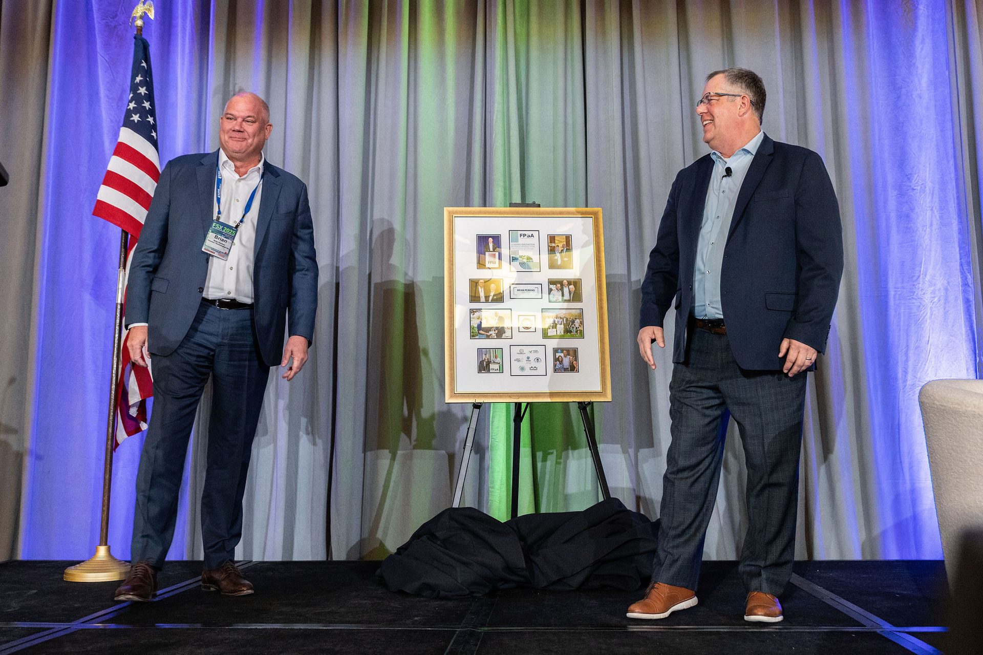Two men stand on a stage with an American flag and a framed award between them.
