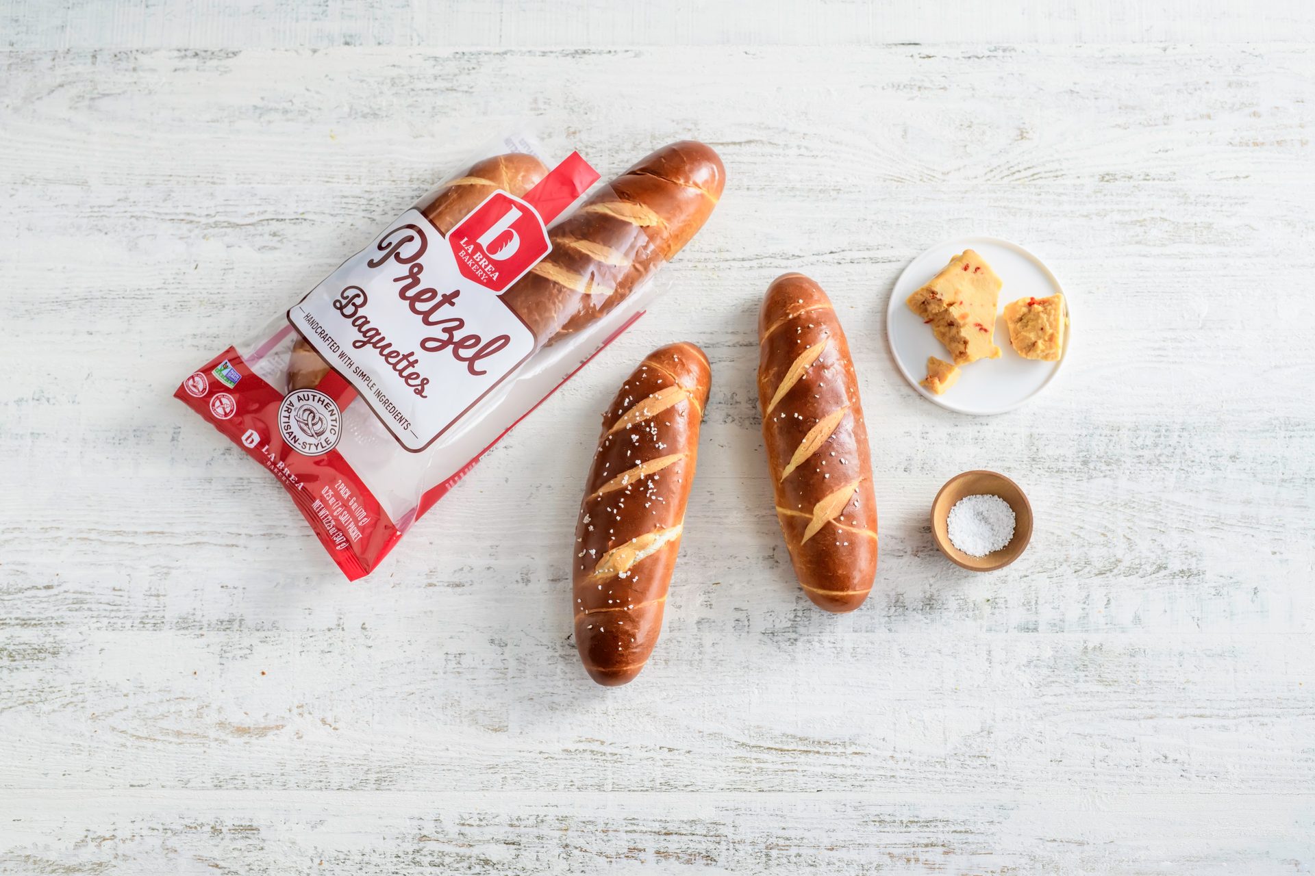 Packaged and loose pretzel baguettes with salt and cheese on a white wooden table.