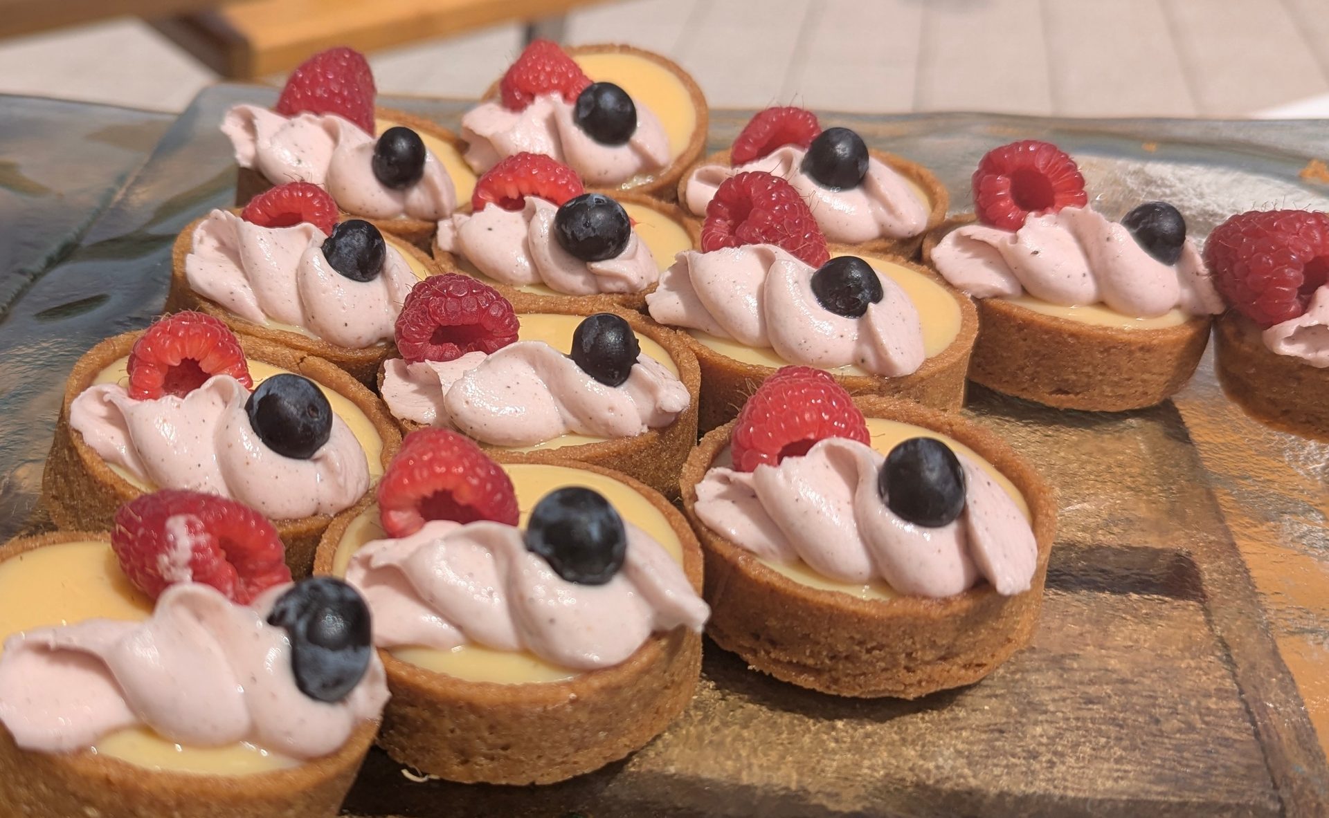 Numerous mini tarts with yellow filling, pink cream, raspberries, and blueberries on a gold tray.