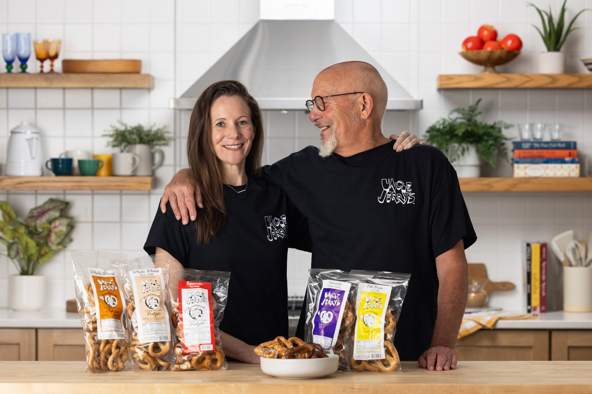 Two smiling people in a kitchen, wearing "Uncle Jerry's" shirts, with bags of pretzels on the counter.