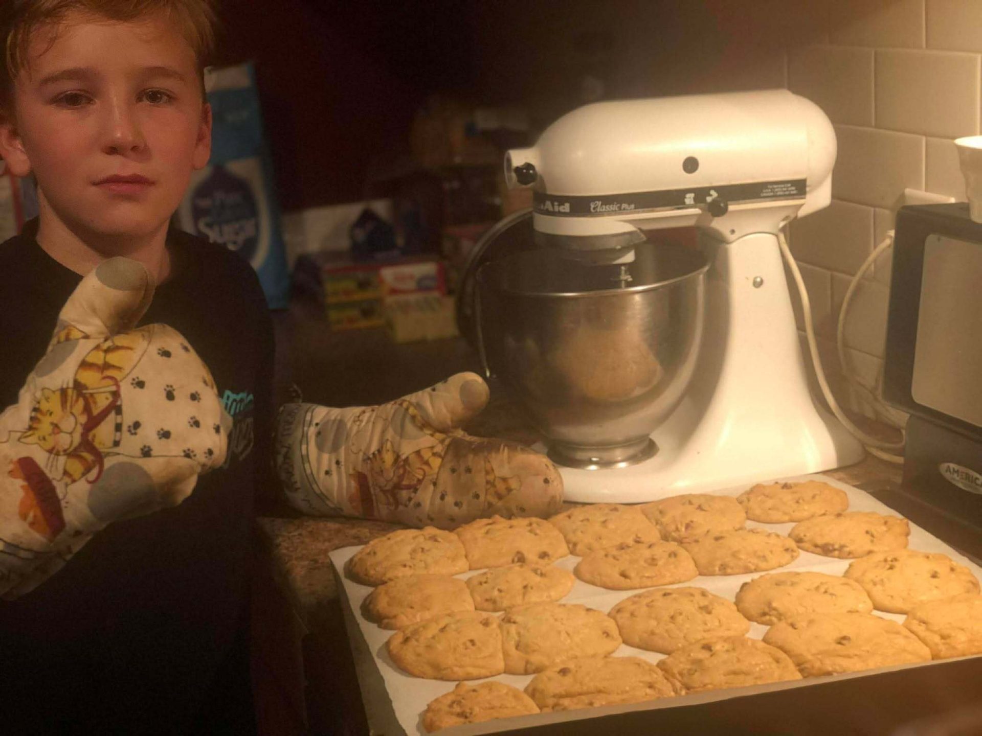 A boy wearing oven mitts proudly displays a tray of freshly baked cookies next to a KitchenAid mixer.