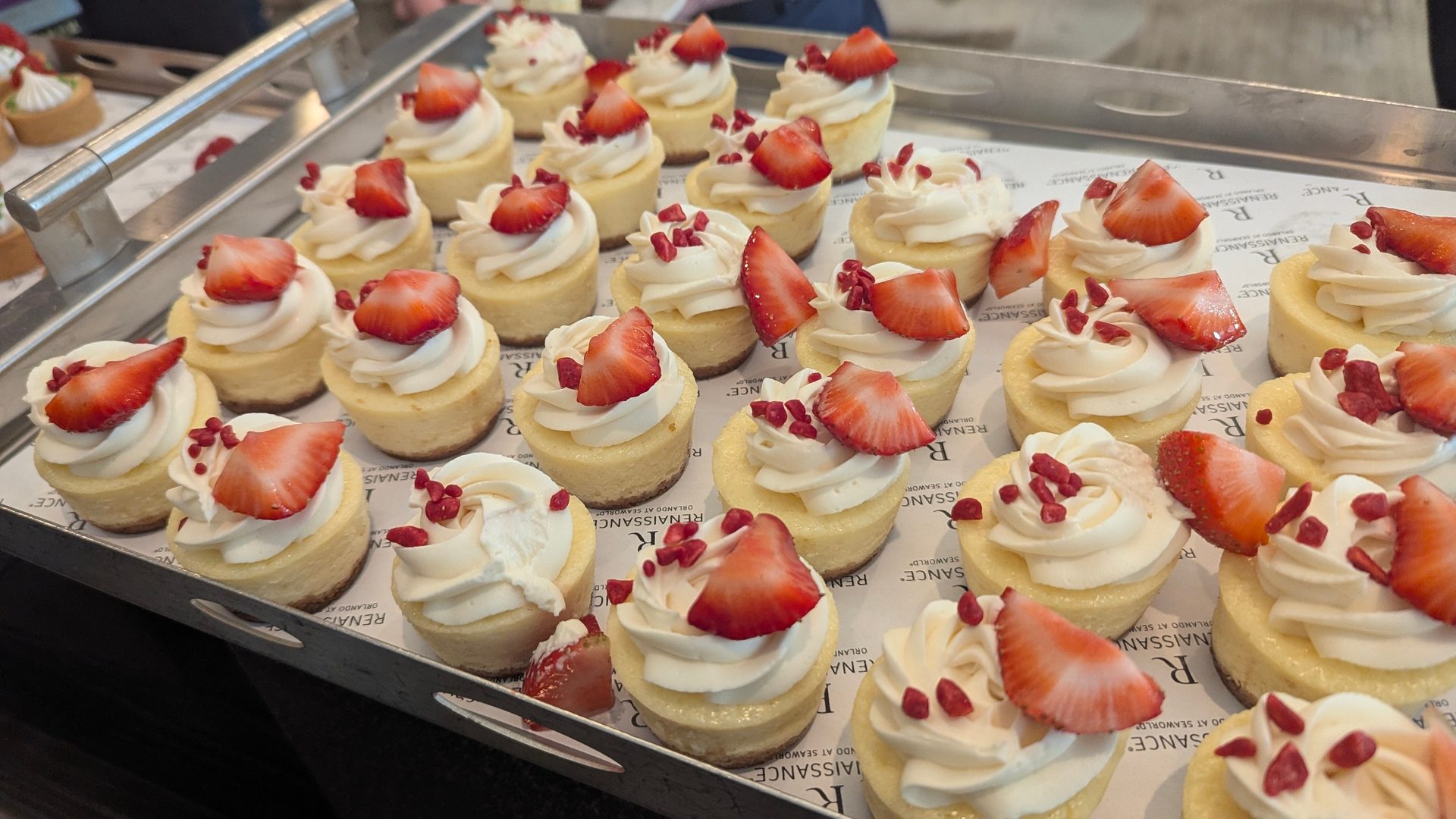 Mini cheesecakes topped with strawberries, white frosting, and red sprinkles on a tray.