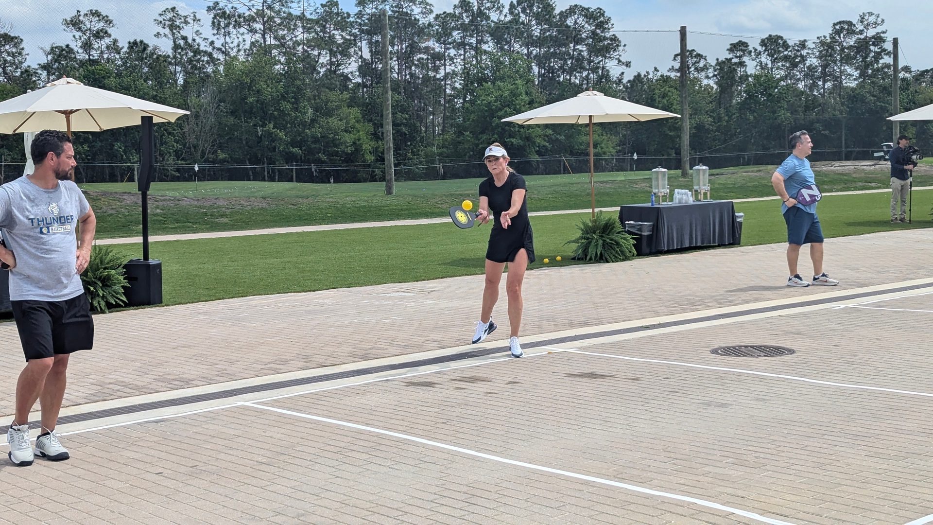 A woman plays pickleball on an outdoor court with two men watching nearby and trees in the background.