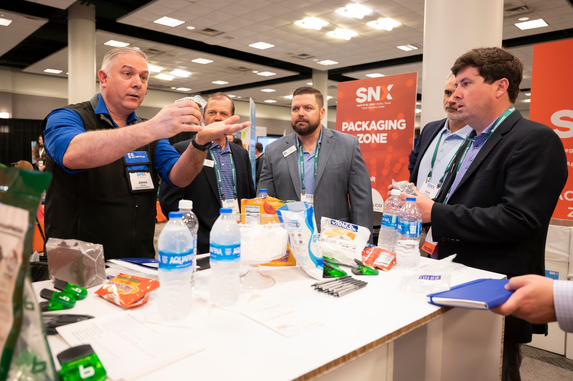 Man presenting items at a trade show booth with packaged products and an 'SNX PACKAGING ZONE' banner.