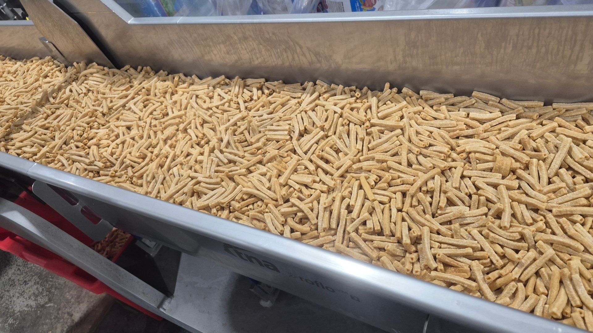A large quantity of light brown, stick-shaped extruded food items moving on a metal conveyor belt.