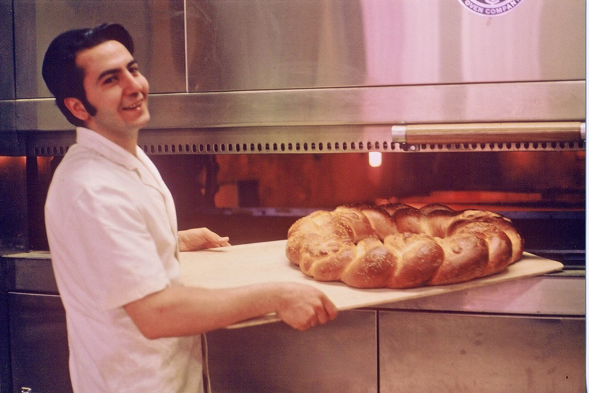 Smiling man holds braided bread on a peel, standing by a hot oven.